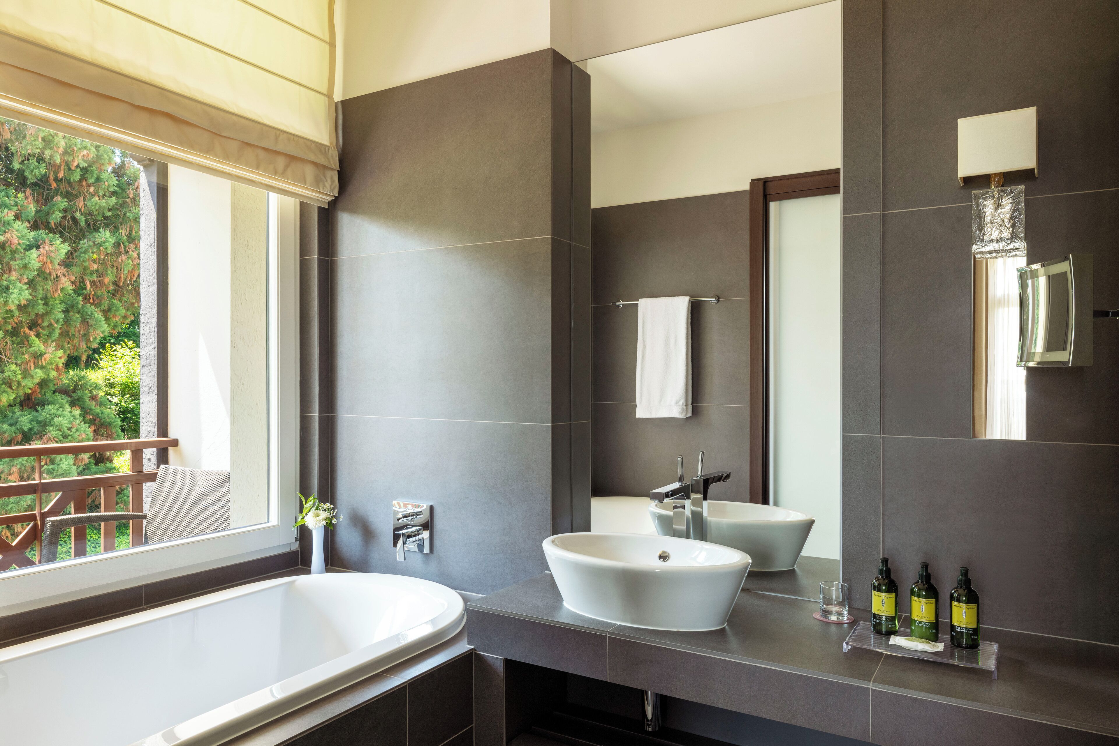 Une salle de bain moderne avec une grande baignoire, un lavabo et un miroir. Une fenêtre offre une vue sur la verdure à l’extérieur.