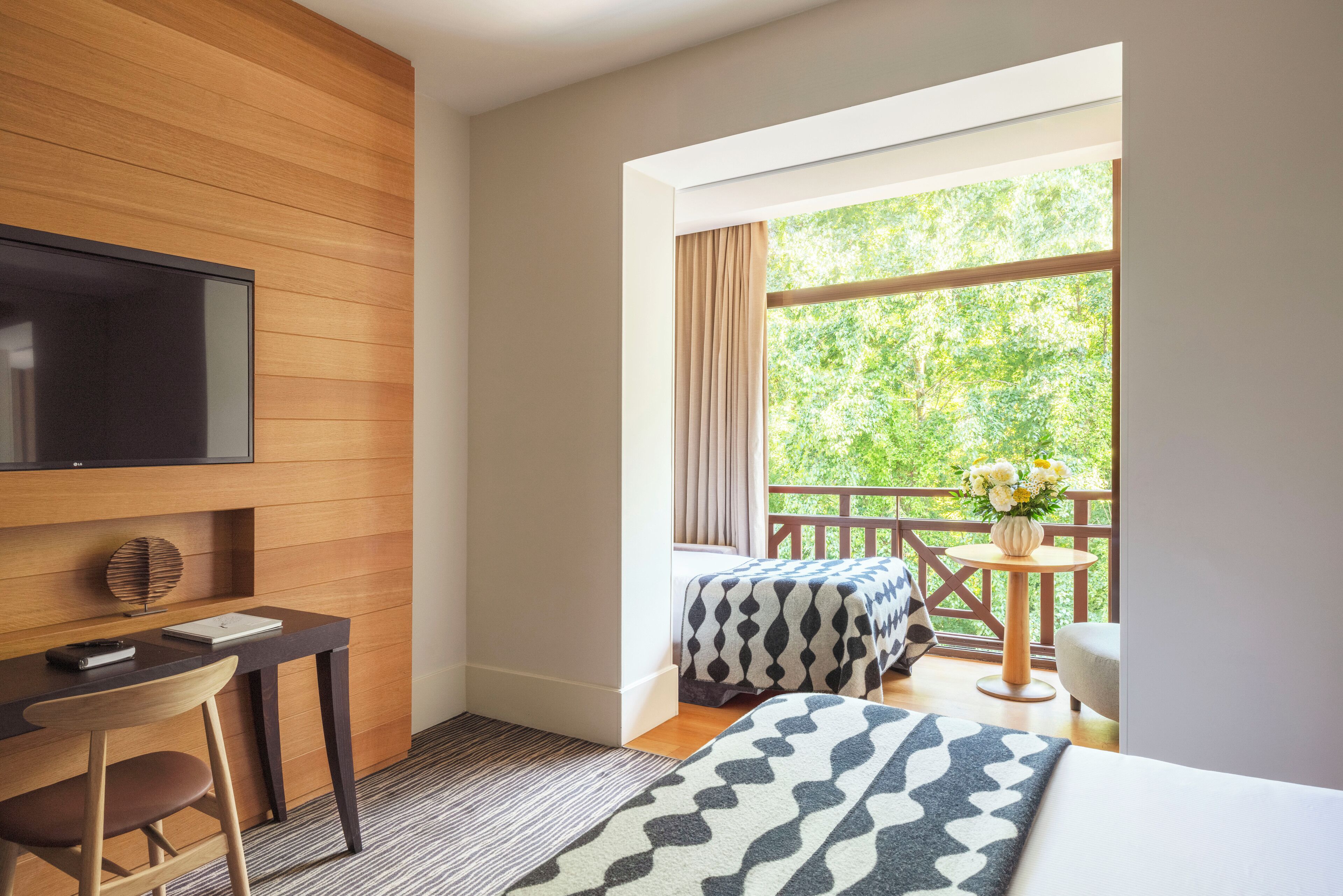 A hotel room with a wooden wall, a flat-screen TV, a desk with a chair, two beds with patterned bedding, a small table with a vase of flowers, and a balcony with a view of greenery.