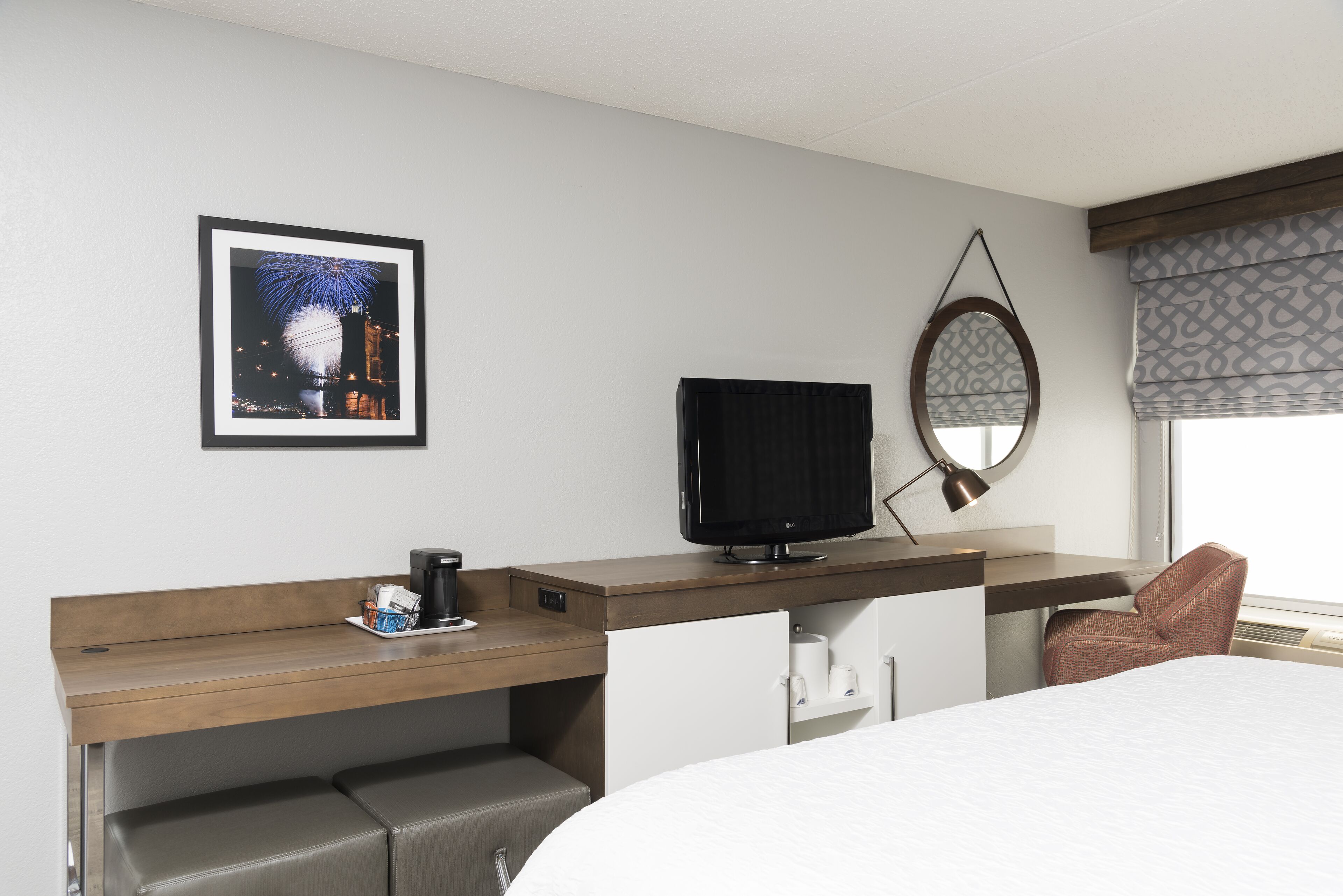 Habitación de hotel con cama, escritorio con televisor, una silla, cafetera y un cuadro en la pared.
