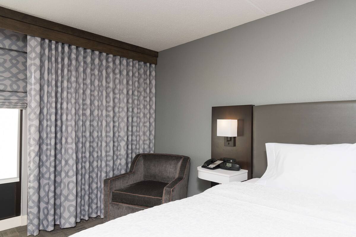 A hotel room with a bed, a chair, a nightstand with a phone, and a wall-mounted lamp.