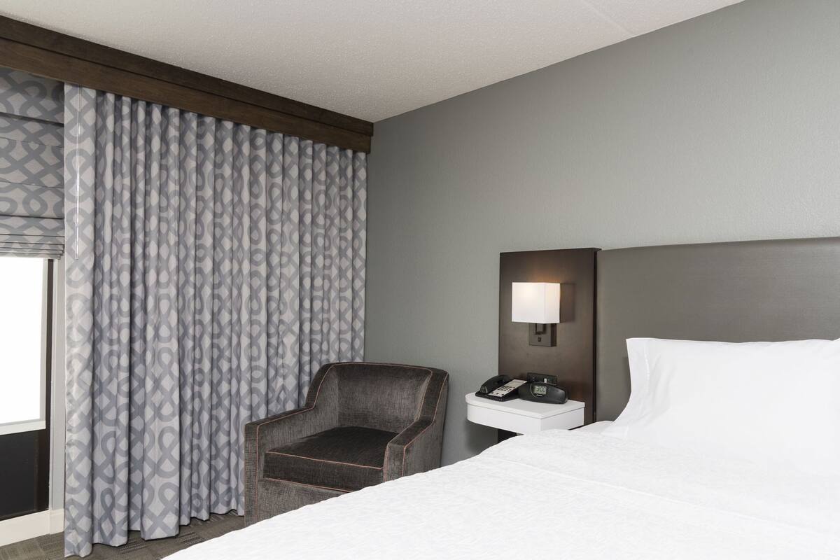 A hotel room with a bed, a chair, a nightstand with a lamp, and a patterned curtain.