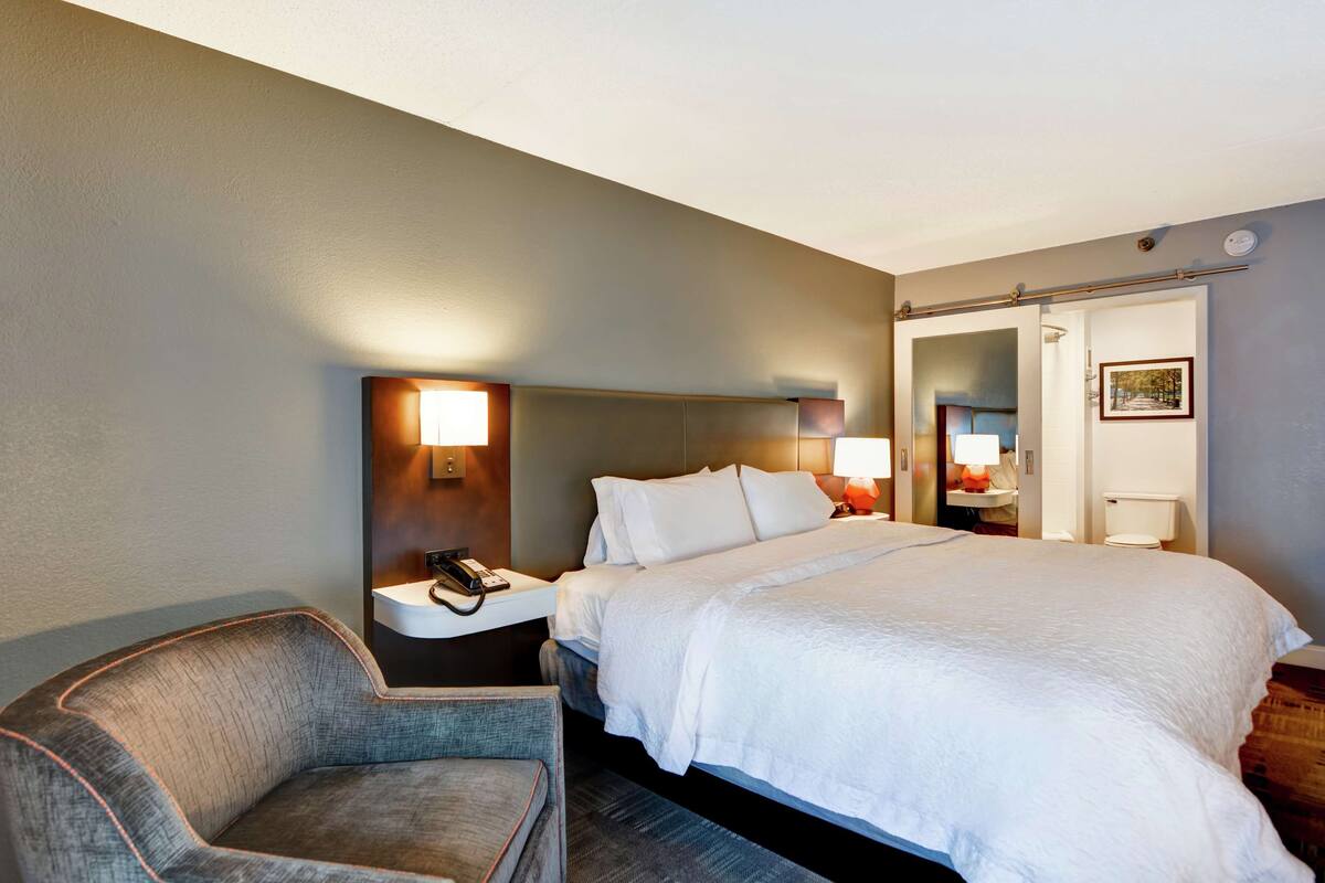 A hotel room with a large bed, a chair, a bedside table with a lamp, and a mirror.