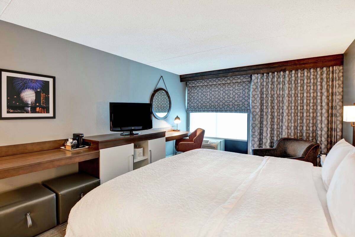 A hotel room with a large bed, a desk with a television, a chair, a small table with a lamp, and a window with curtains.
