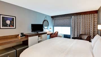 Ein Hotelzimmer mit einem großen Bett, einem Schreibtisch mit Fernseher, einem Sessel, einem kleinen Tisch mit Lampe und einem Fenster mit Vorhängen.