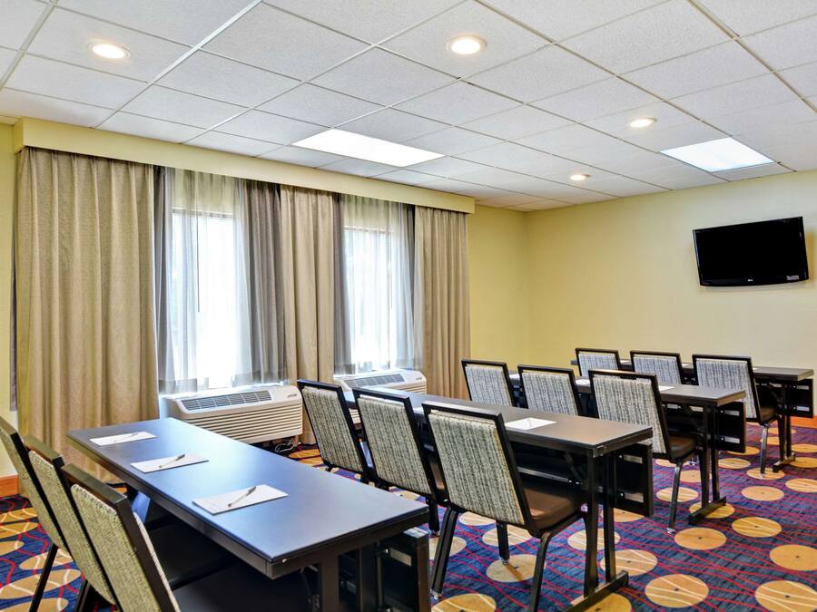 A conference room with tables and chairs, air conditioning units, a flat-screen TV, and large windows with curtains.