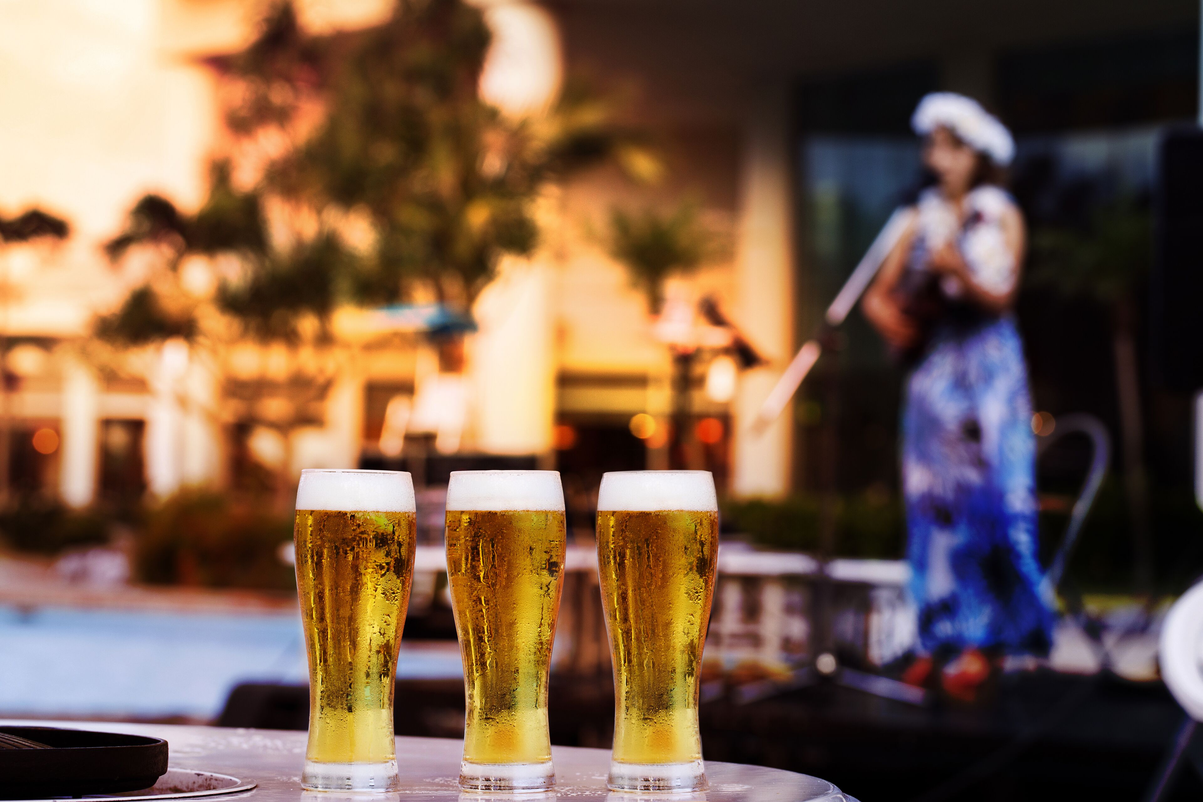 Three glasses of beer on a table with a pool and a statue in the background.