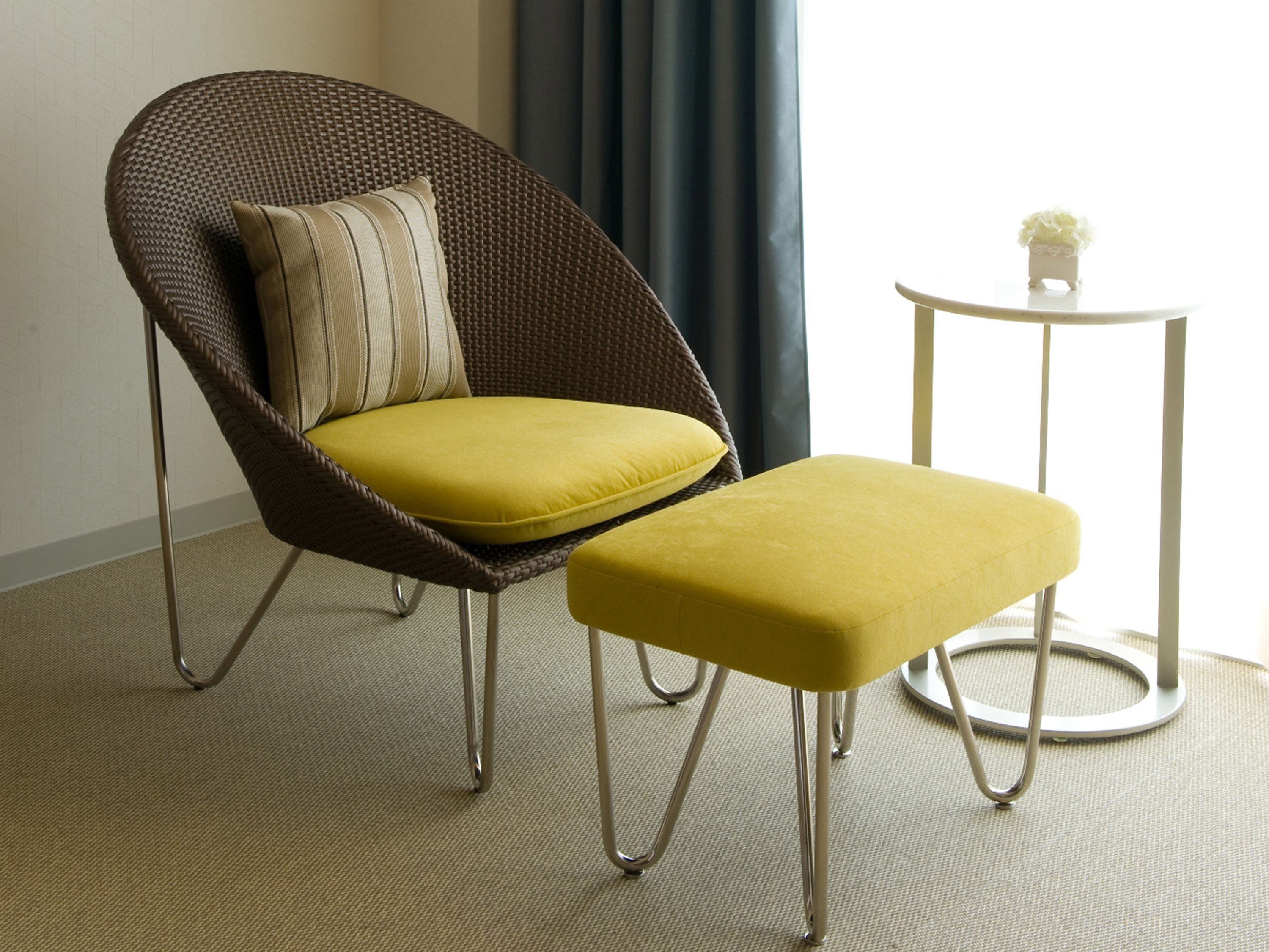 Una silla de mimbre con un cojín amarillo y un reposapiés a juego, una mesita redonda con un jarrón blanco, y vistas de un edificio a través de una ventana con cortina.