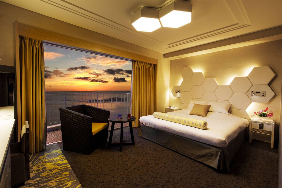 Une chambre d’hôtel avec un lit, une petite table, une chaise, une table de chevet et une vue sur le coucher de soleil par la fenêtre.
