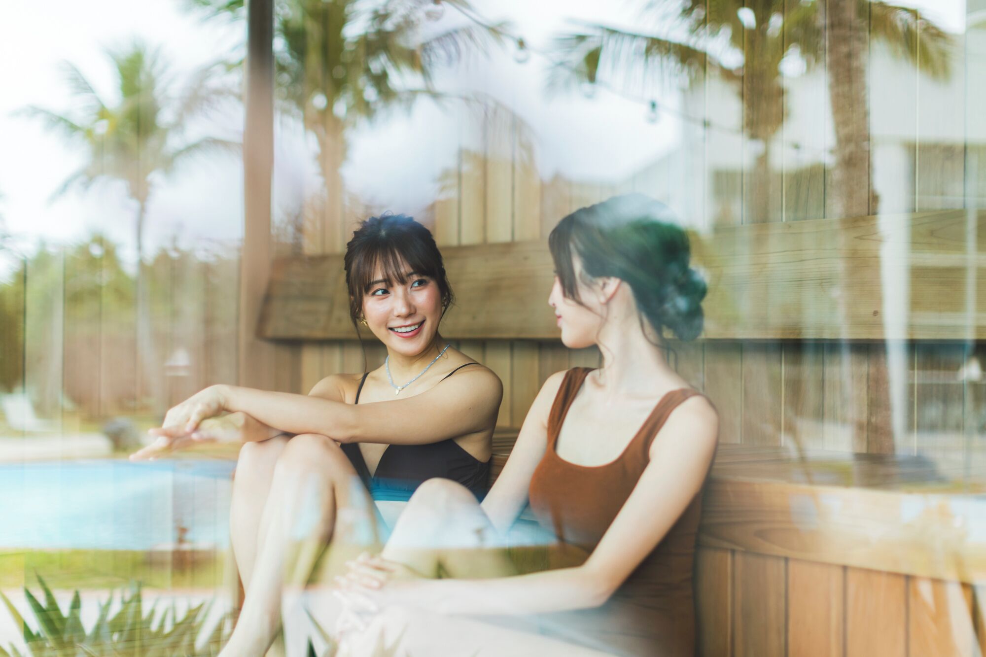 Dos personas sentadas junto a una piscina, con estructuras de madera y palmeras al fondo.