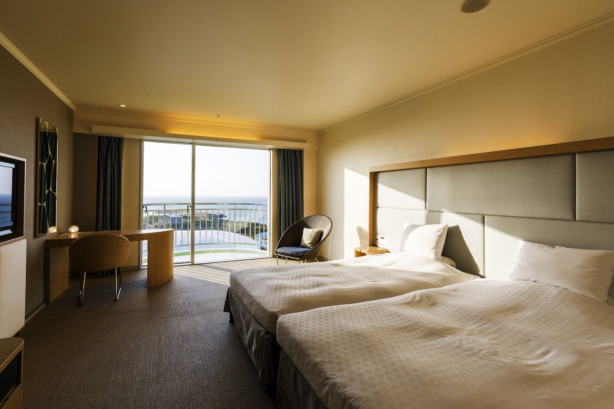Une chambre d’hôtel avec deux lits, un bureau, une chaise et une grande fenêtre donnant sur un beau paysage.
