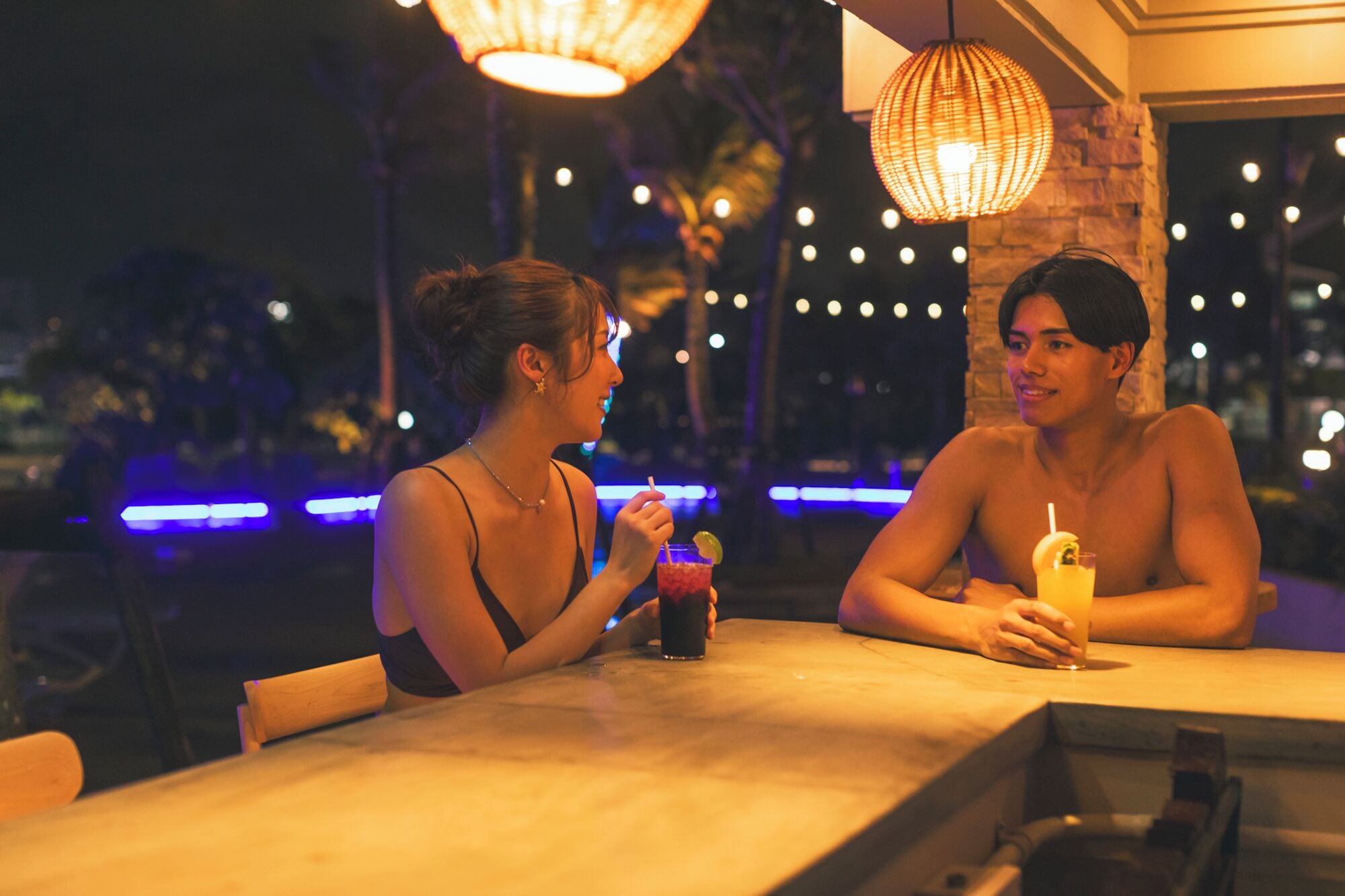 Dos personas sentadas en una mesa con bebidas, en un ambiente interior tenuemente iluminado con luces colgantes.