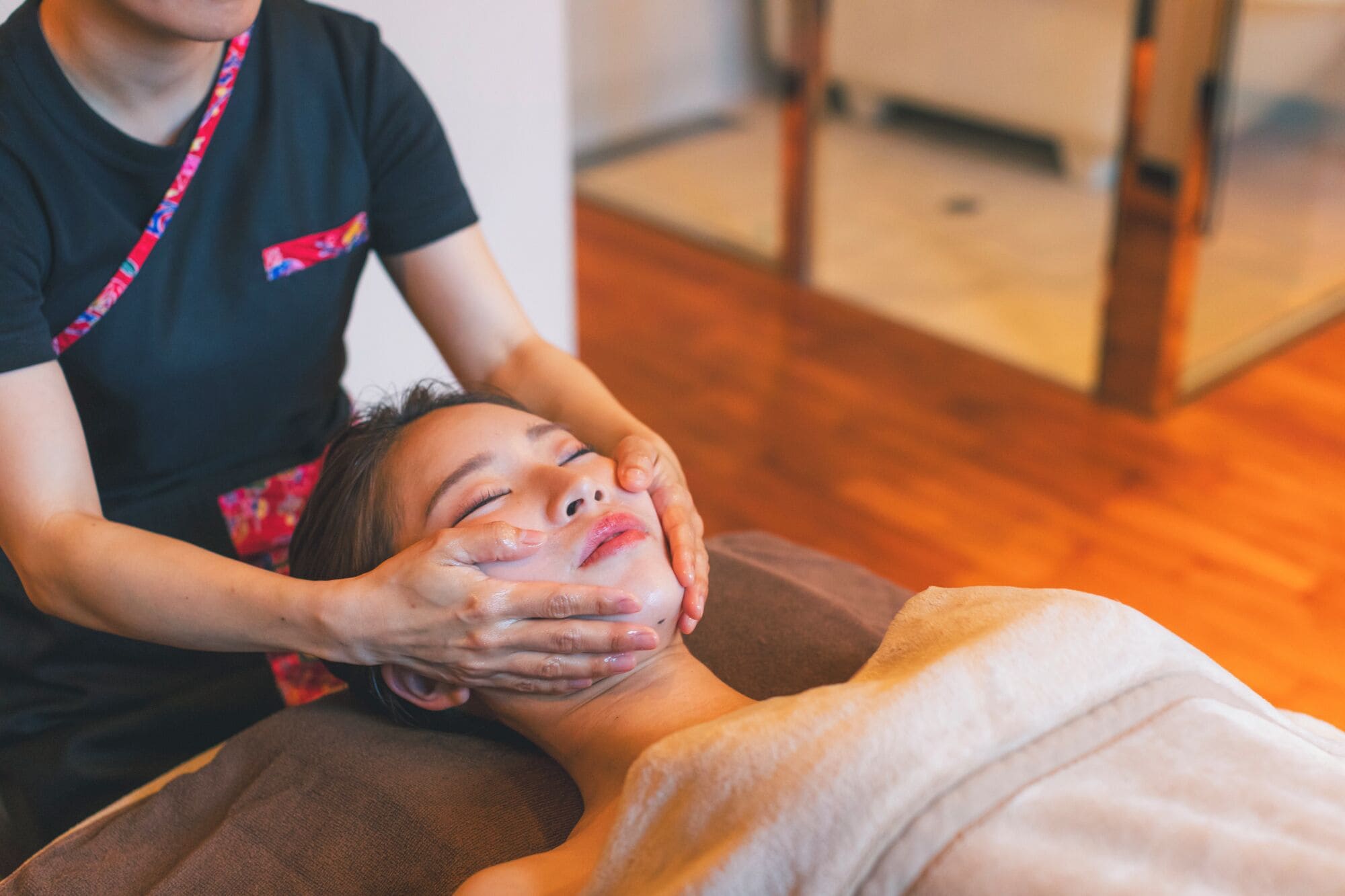 A person receiving a facial treatment in a room with wooden flooring and a visible chair.