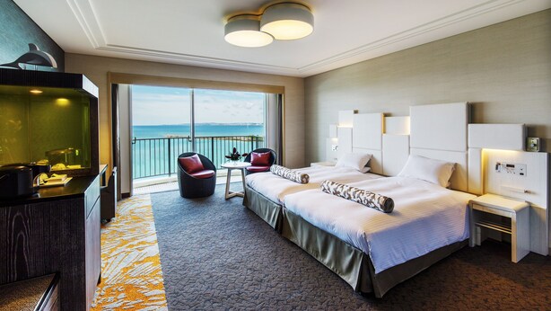 A hotel room with a large bed, a desk, a chair, a small table, and a view of the sea. - Laguna Garden Hotel (Ginowan)
