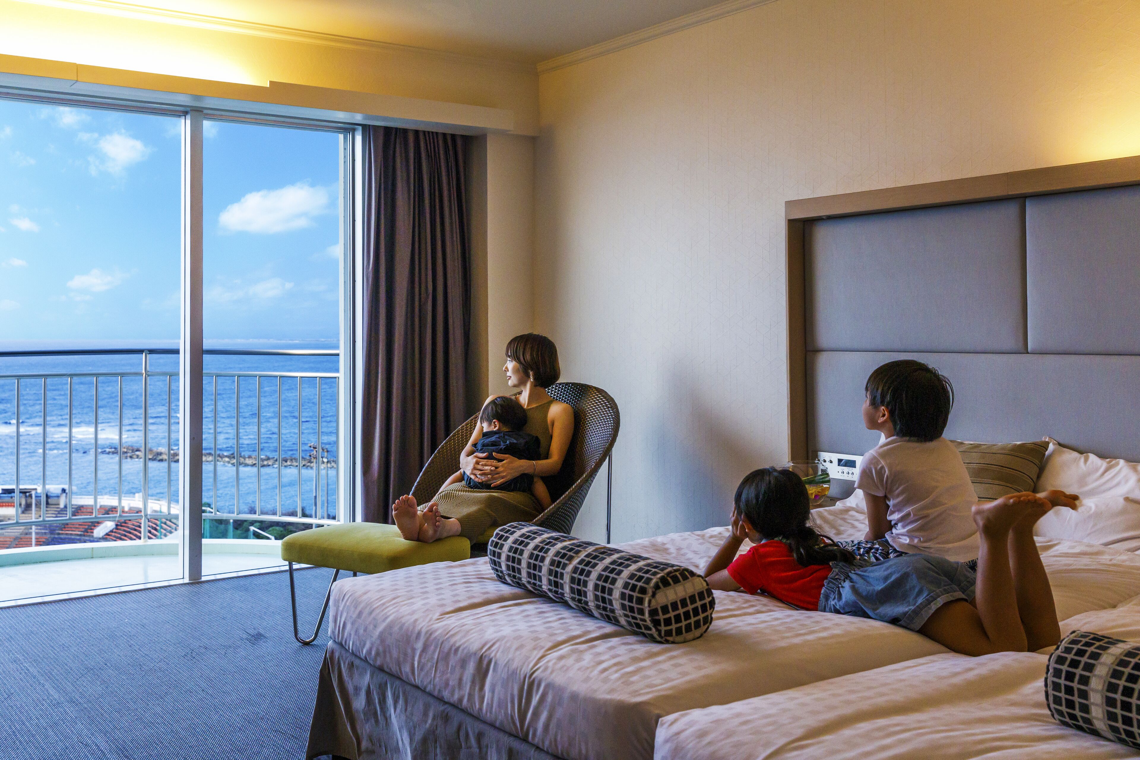 A hotel room with a large window overlooking the sea, a bed with pillows, and a chair.