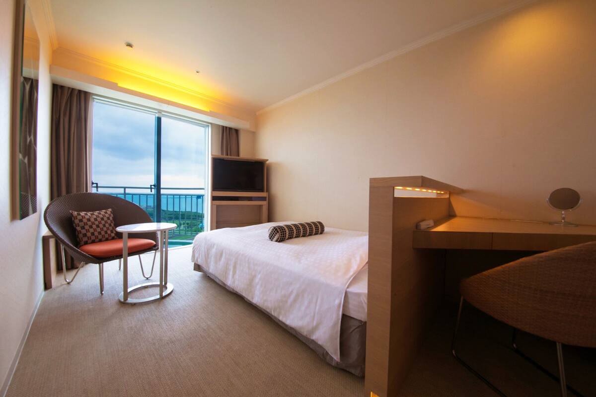 Une chambre d’hôtel avec un lit, une chaise, une petite table, un téléviseur et un balcon avec des rideaux.