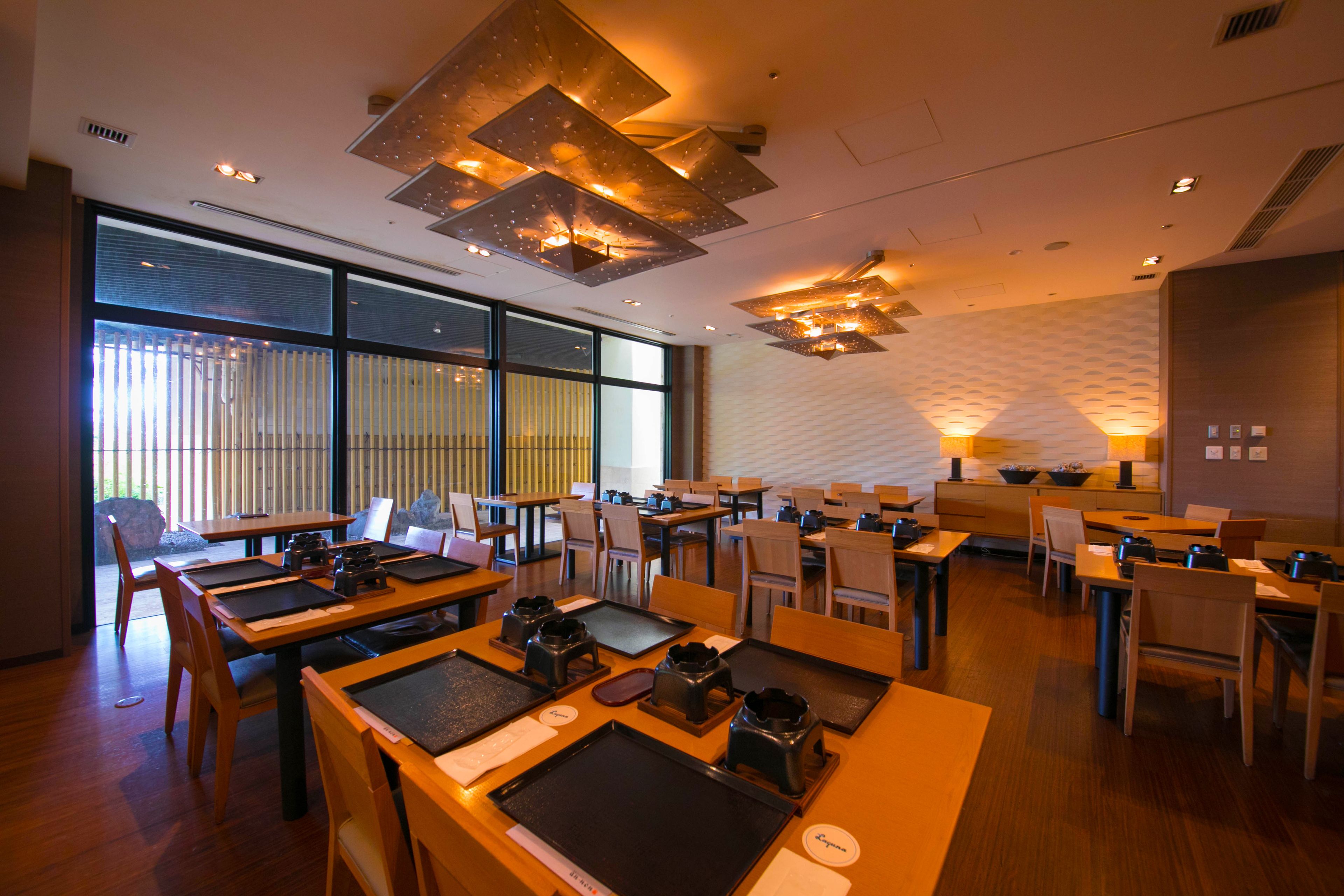 Un restaurante moderno con varias mesas y sillas, ventanas amplias y lámparas de techo contemporáneas.