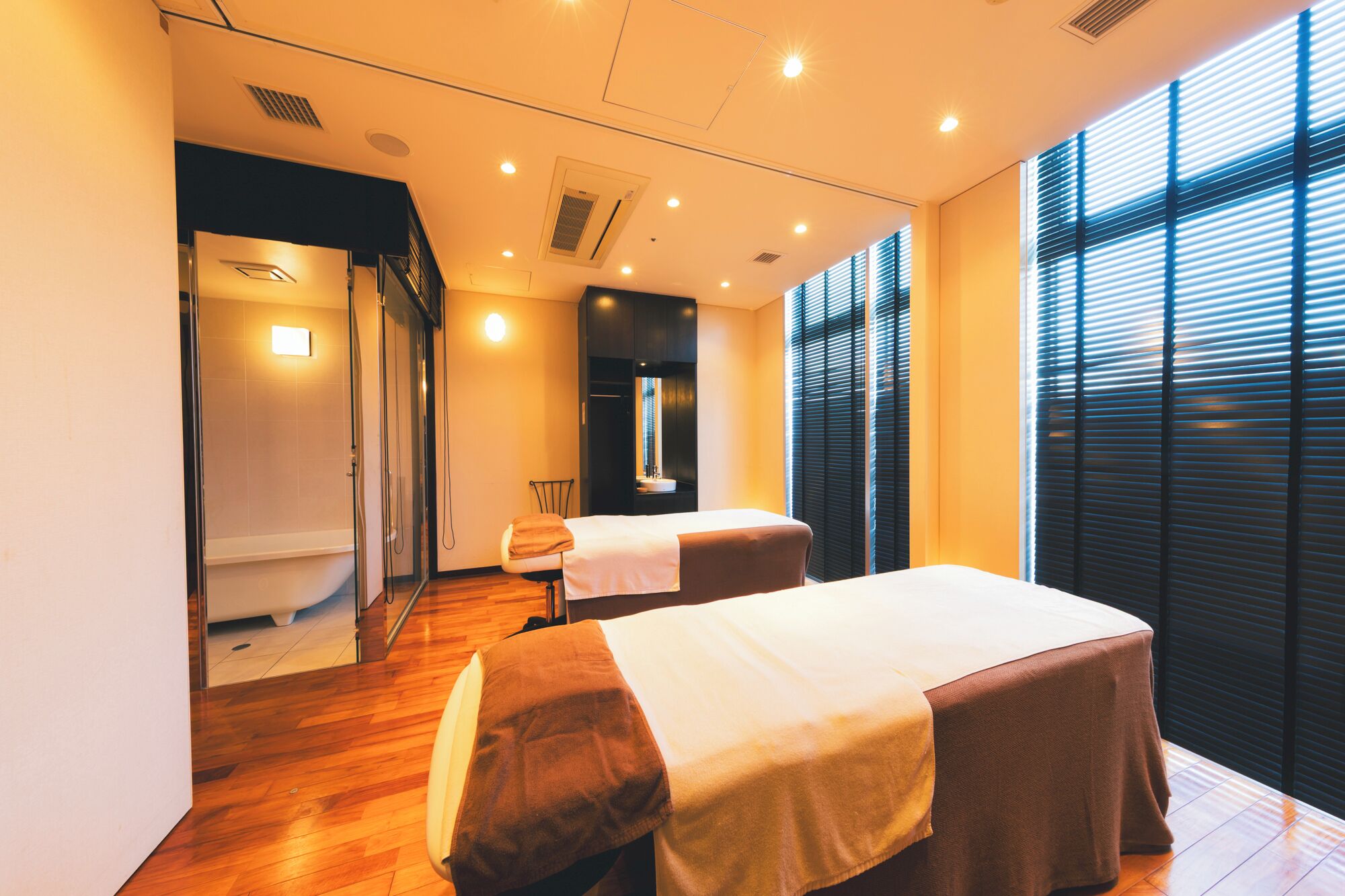 A room with a wooden floor, two massage tables, a small chair, a mirror, and a window with blinds.
