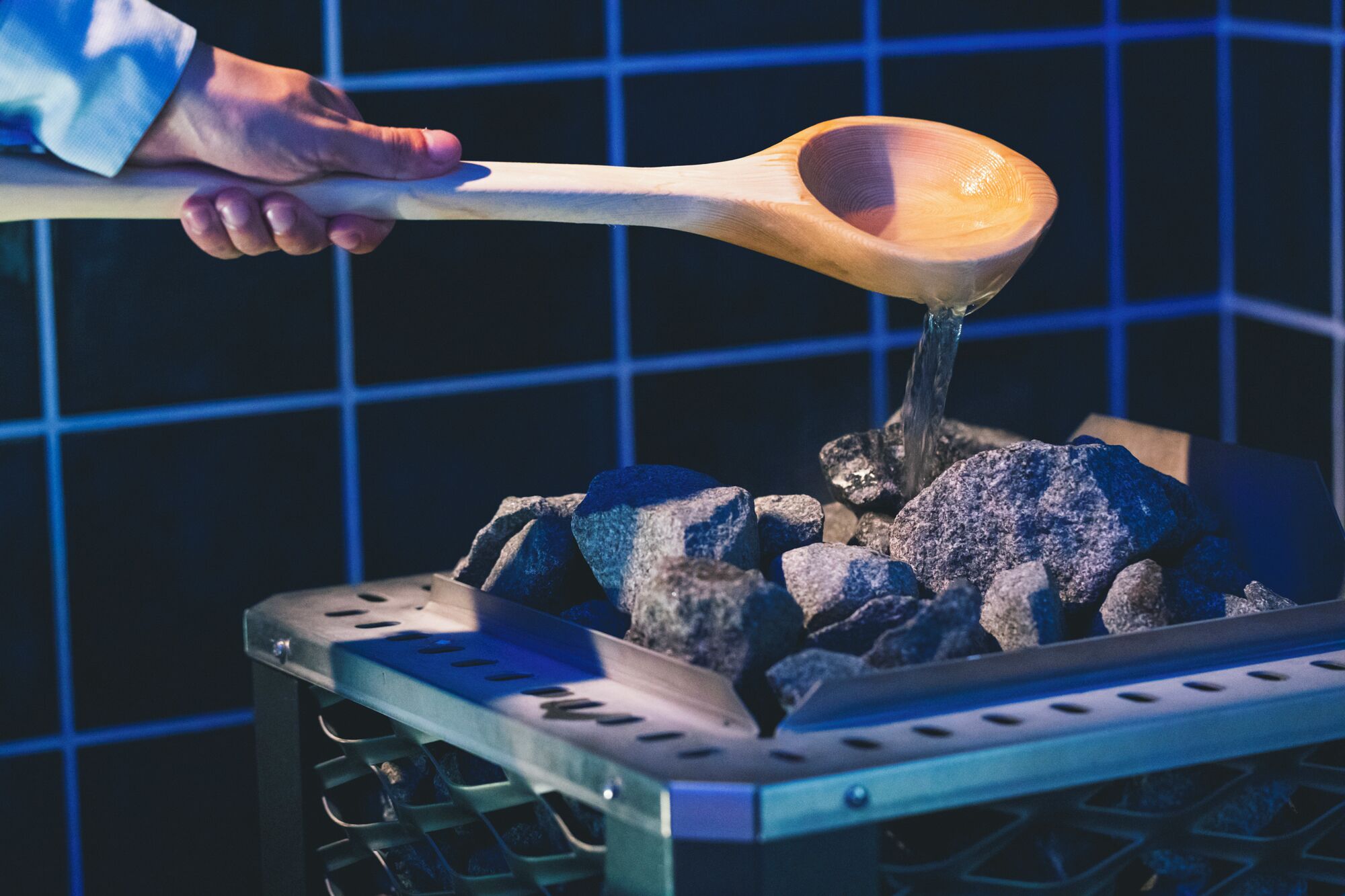 A hand holding a wooden ladle pouring liquid over charcoal in a metal container.
