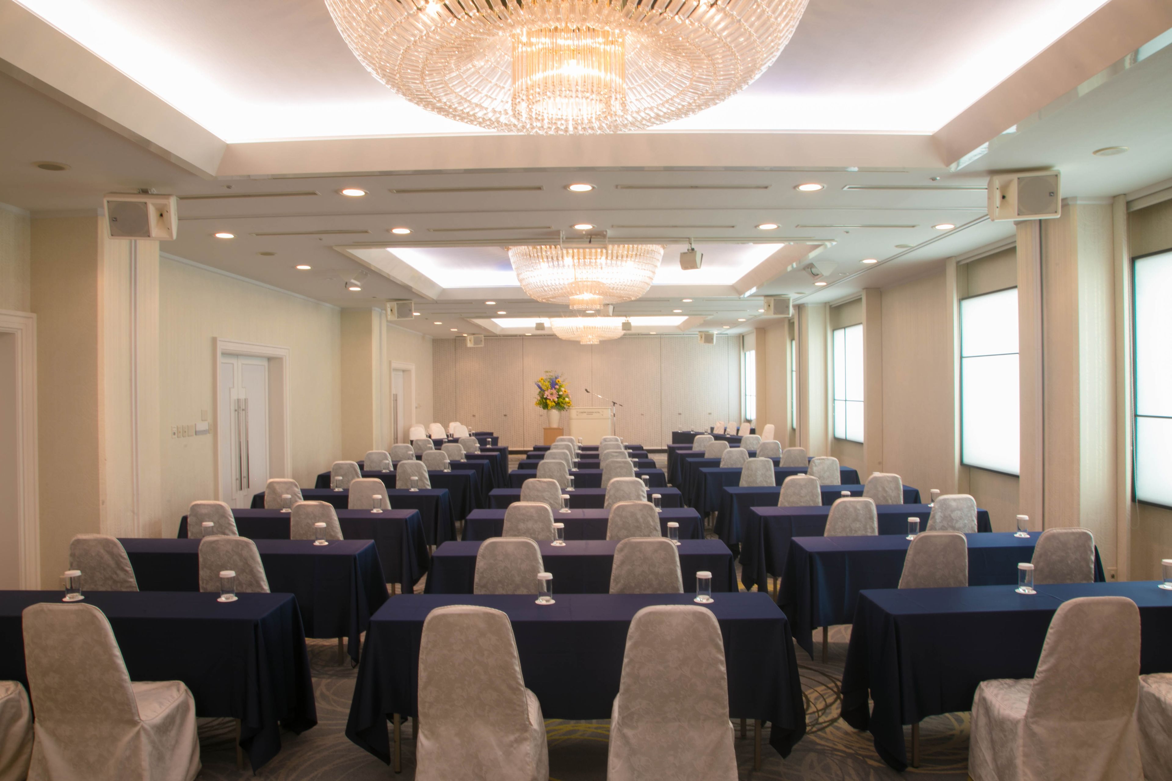Une salle de conférence avec des rangées de tables et de chaises, un grand lustre et un pupitre agrémenté d’un arrangement floral.