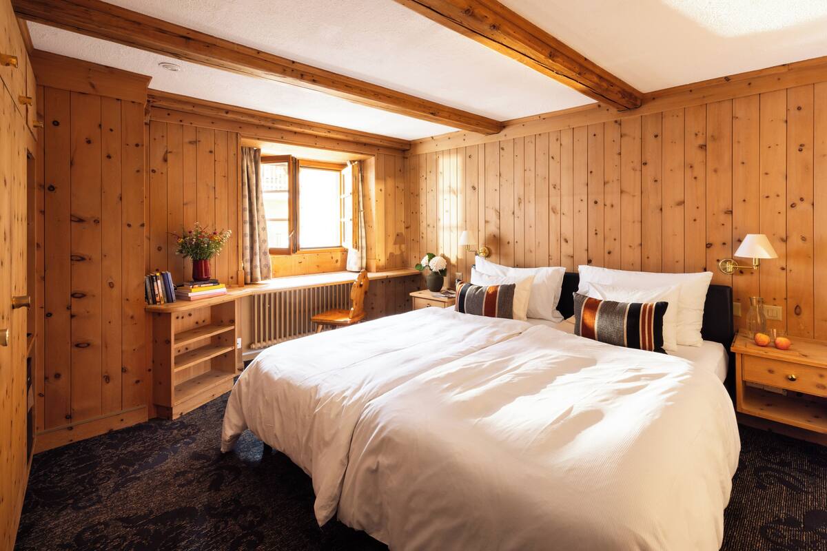 A wooden-paneled bedroom with a large bed, a nightstand, a window with curtains, and a wooden chair.