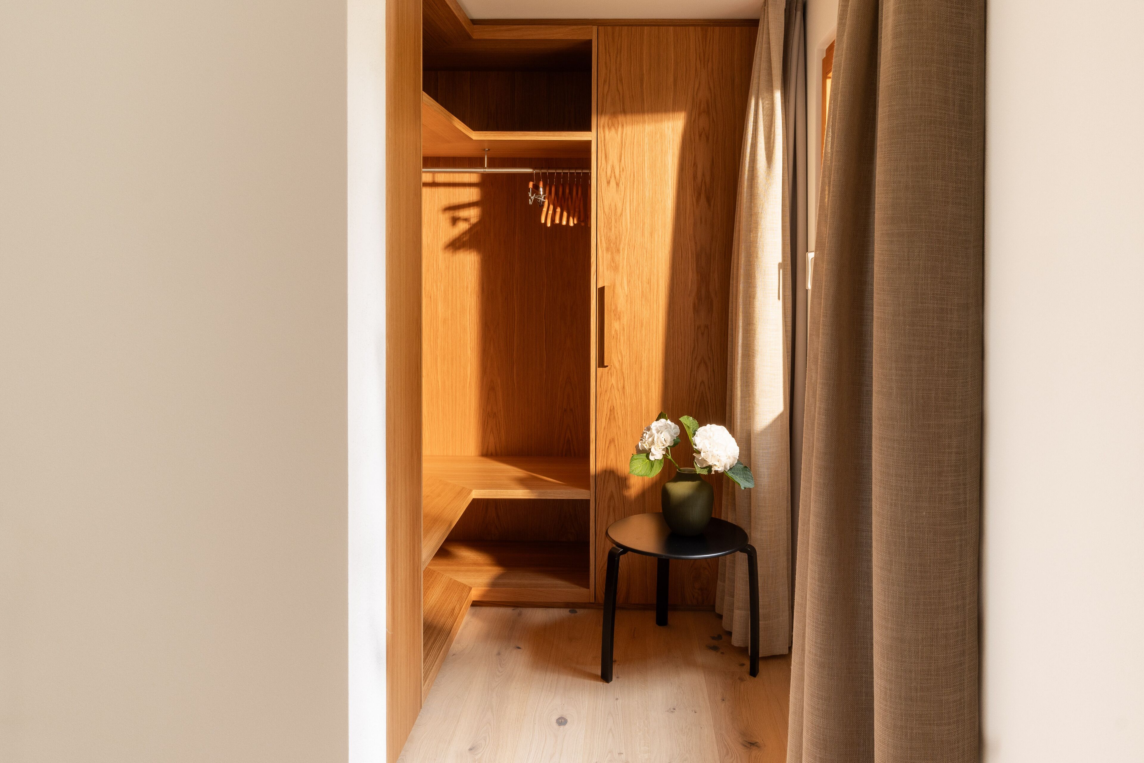A wooden closet with a hanging rack, a small table with a vase of flowers, and a curtain partially drawn to one side.