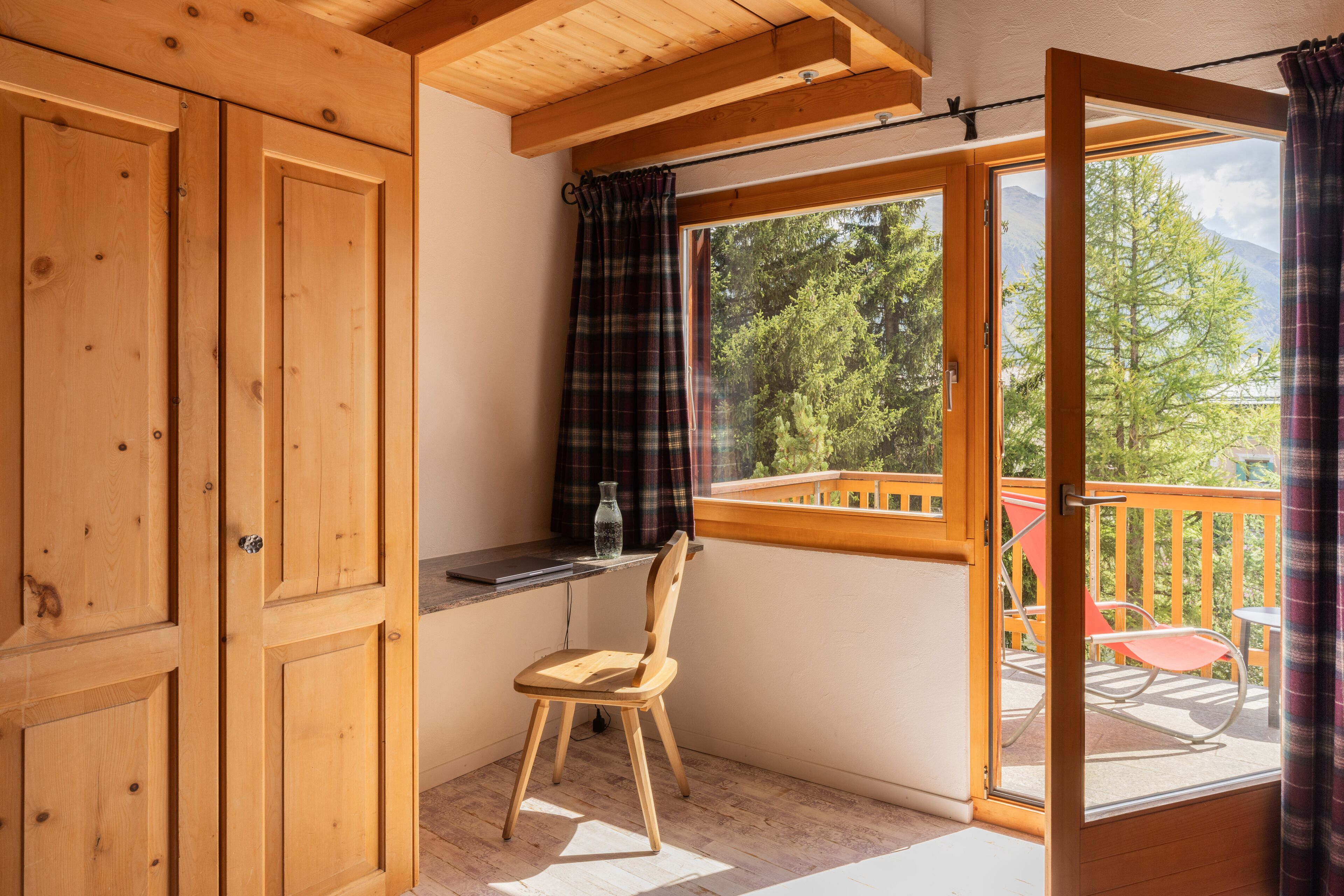 A wooden balcony with a view of trees and mountains, a chair, and a small table.