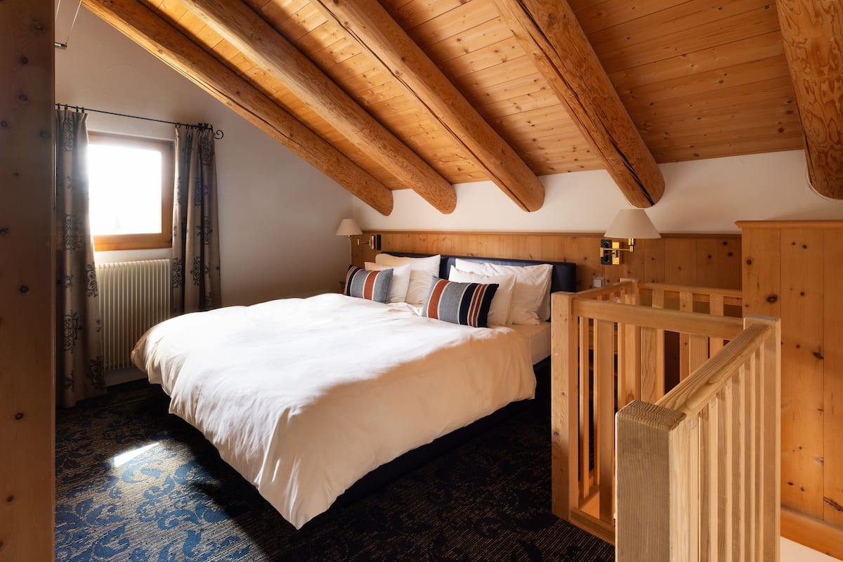 A bedroom with a large bed, wooden ceiling beams, a radiator, and a window with curtains.