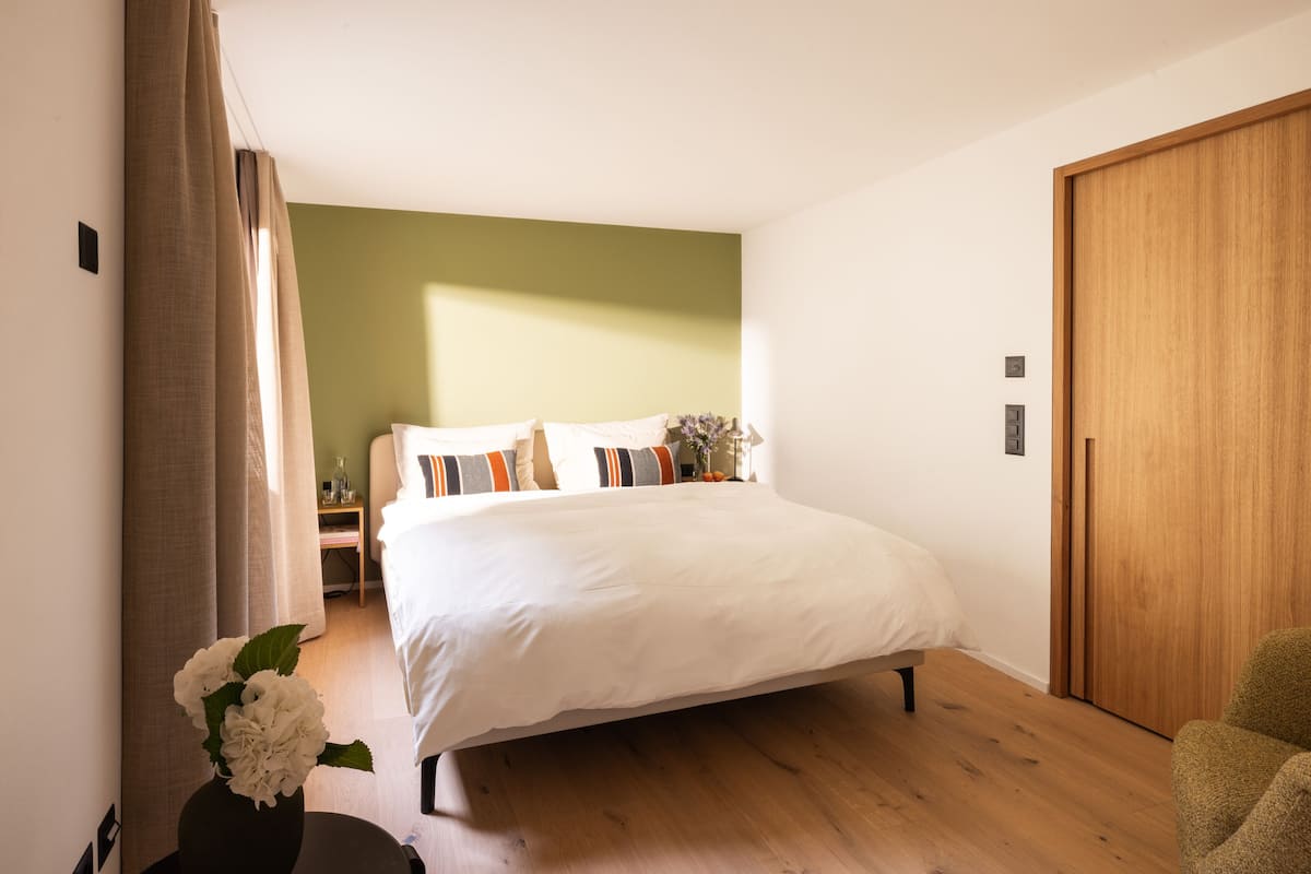A bedroom with a large bed, wooden flooring, a green accent wall, a wooden door, and a nightstand with a lamp and vase.
