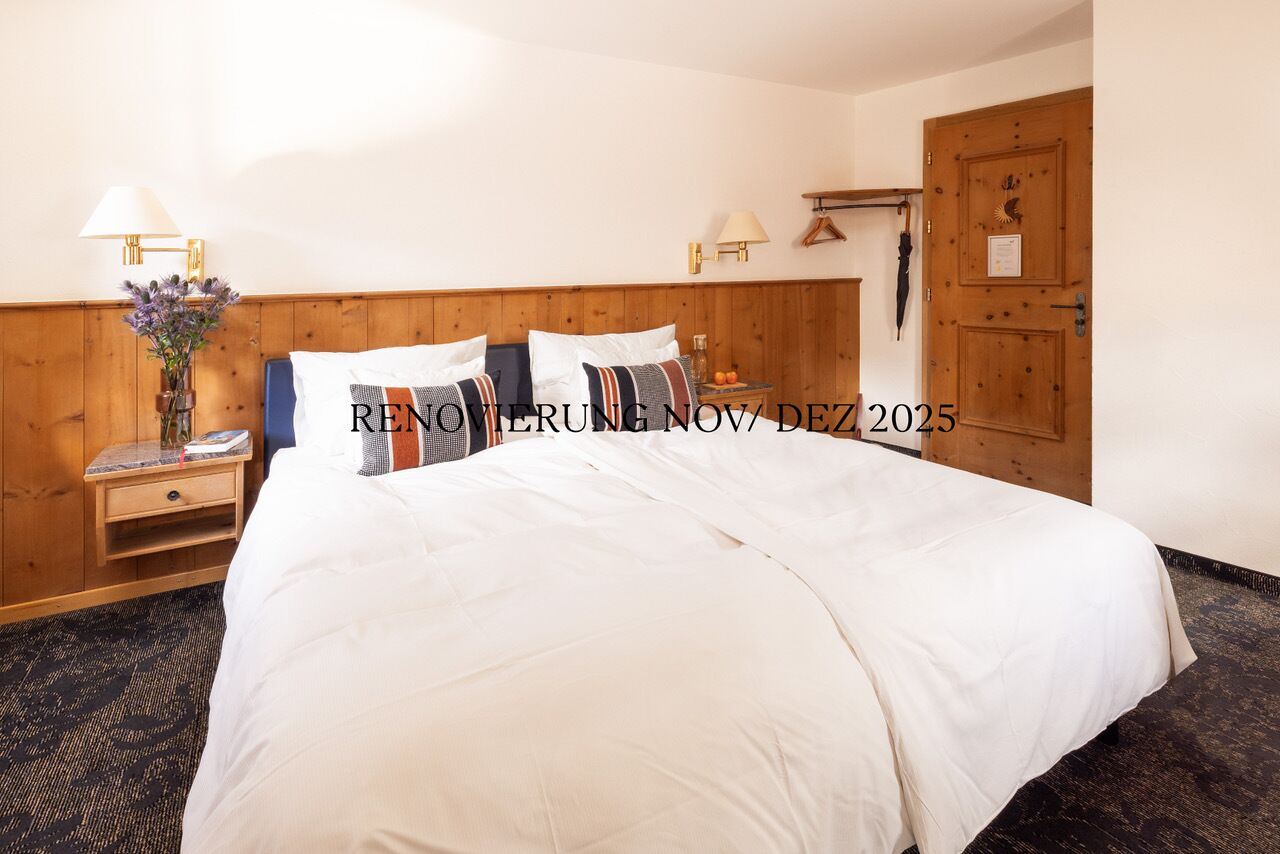 A hotel room with a large bed, wooden headboard, two bedside tables with lamps, a small table with a vase of flowers, and a wooden wardrobe.