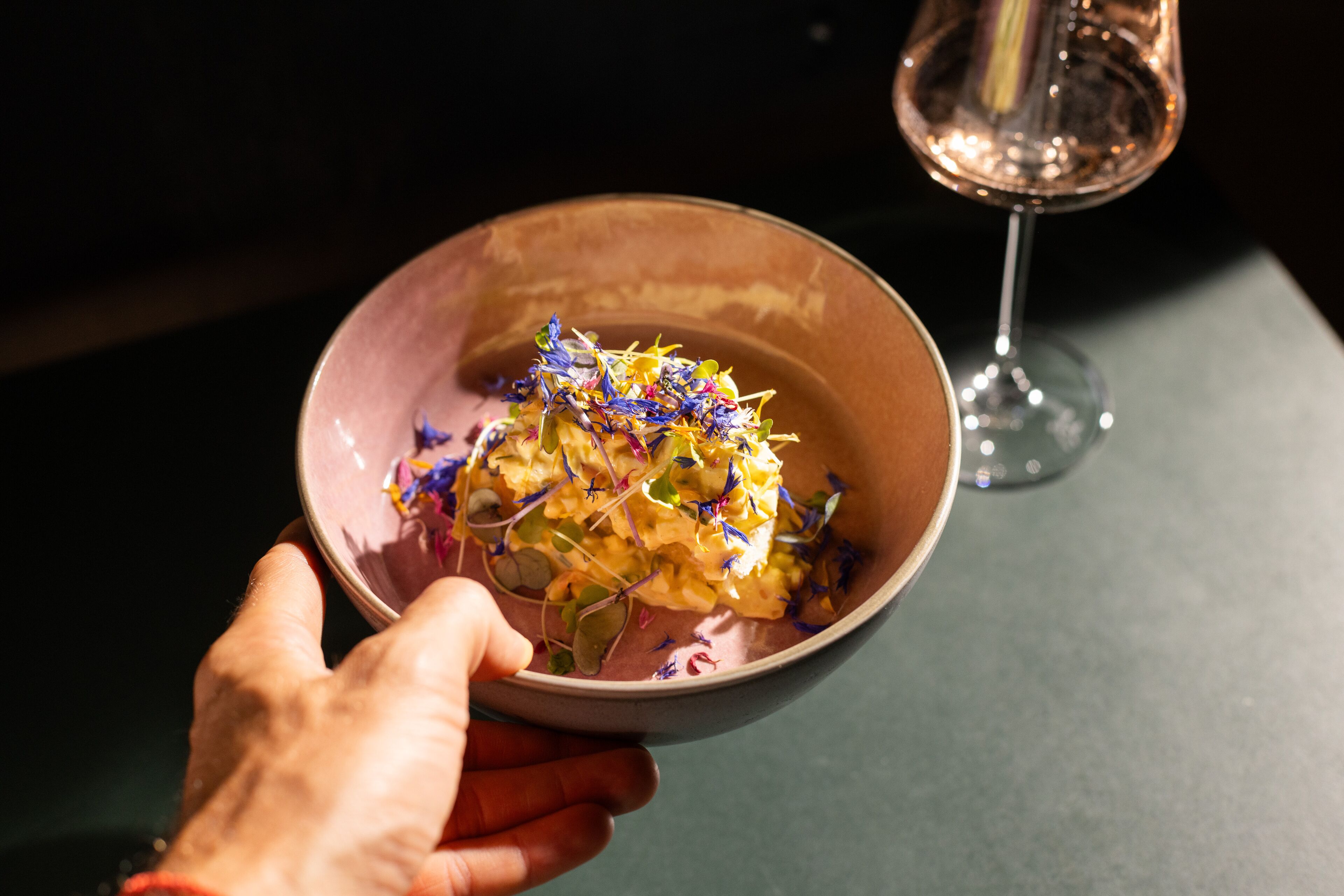 A hand holding a bowl of food with a glass of wine in the background.