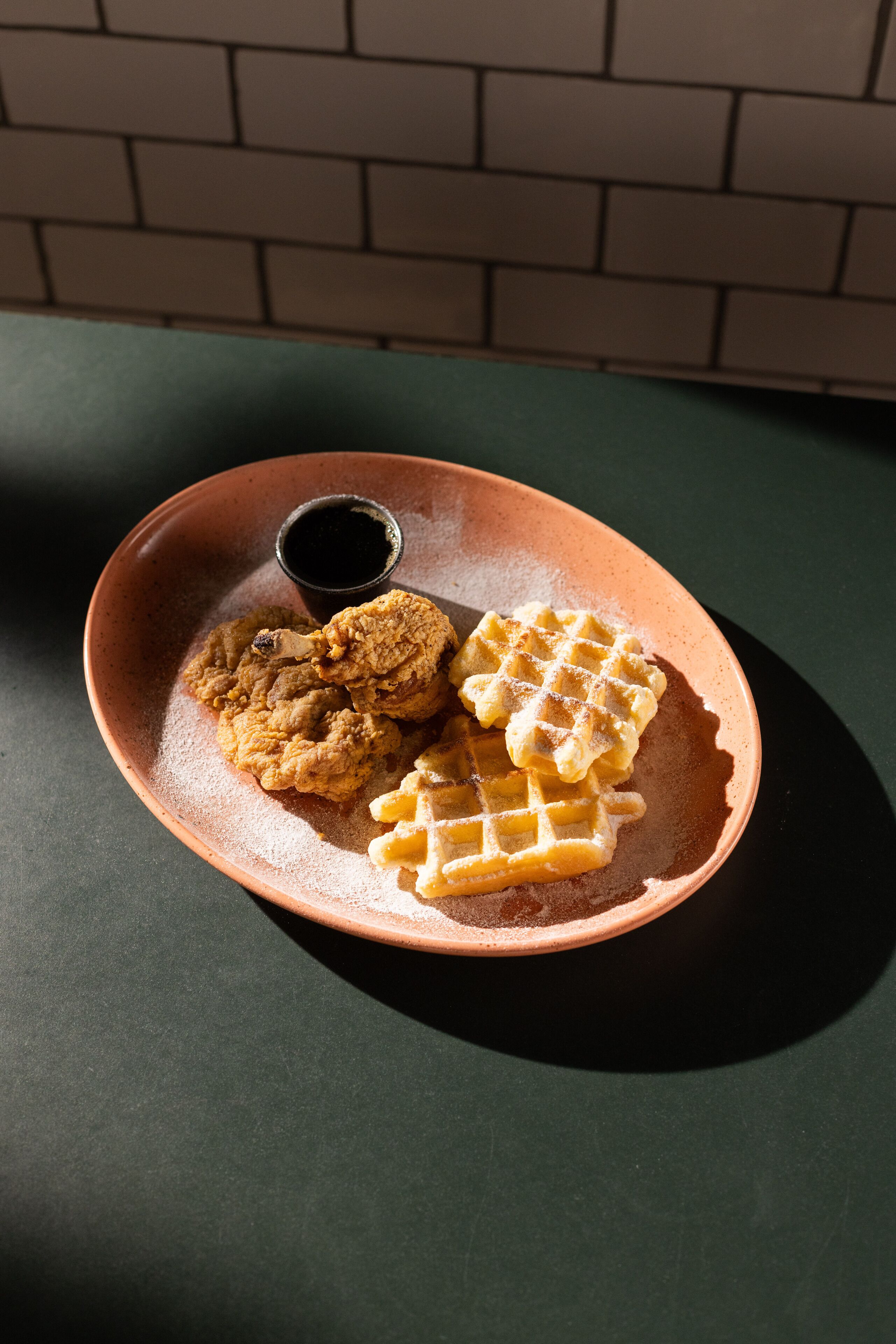 A plate with fried chicken, waffles, and a small cup of sauce on a dark surface.