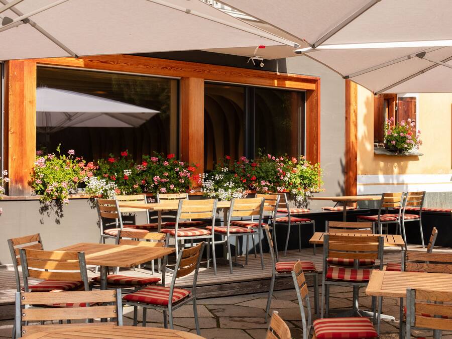 An outdoor cafe with wooden tables and chairs, large windows, and hanging flower baskets.