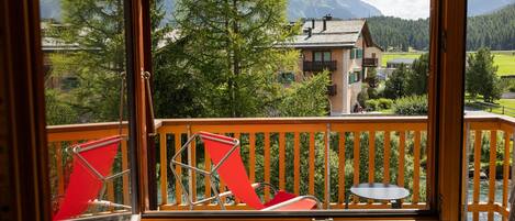A balcony with a view of mountains, a wooden railing, and two red chairs.