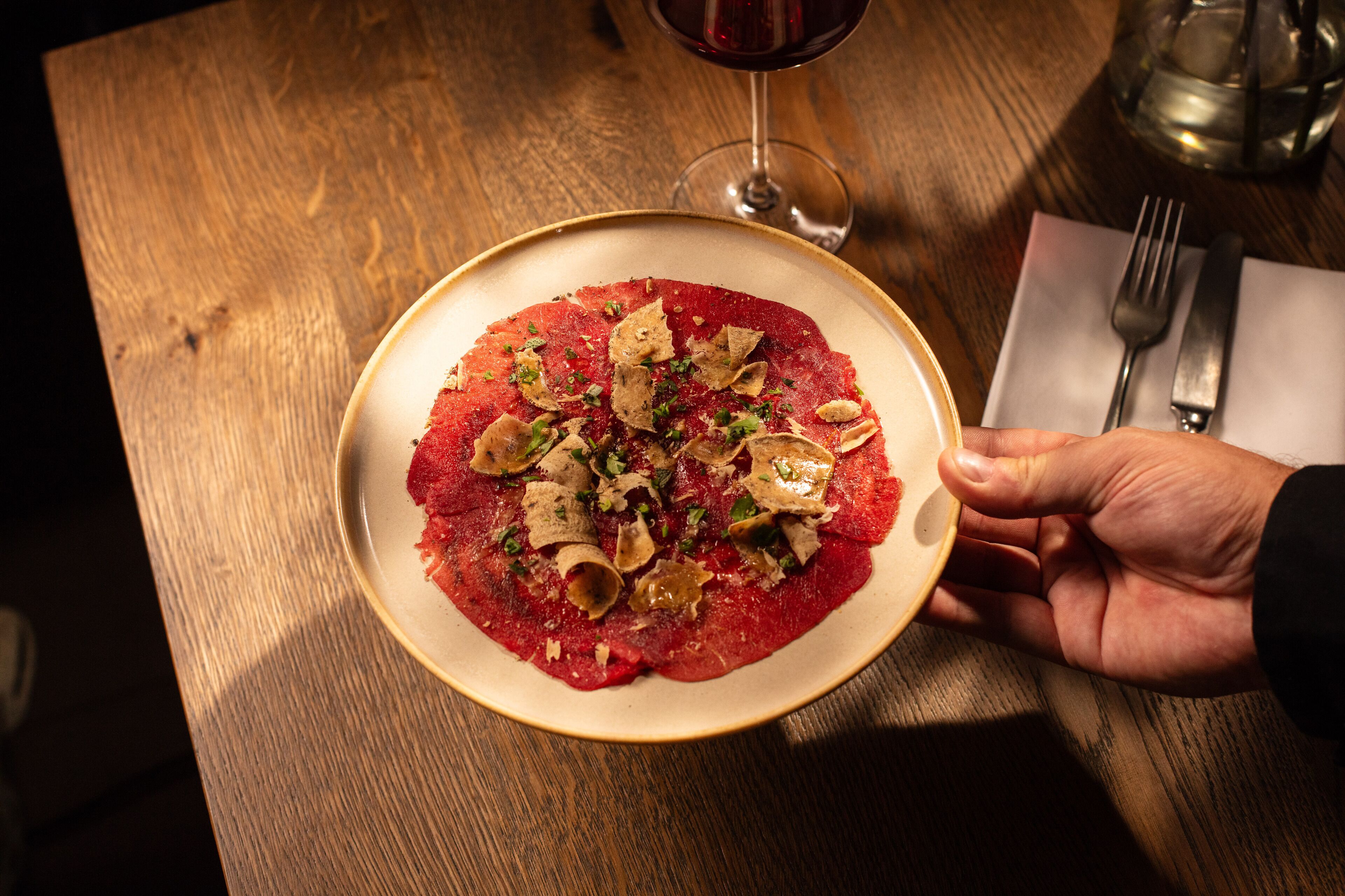 Un plato de comida sobre una mesa de madera con una copa de vino tinto y cubiertos.