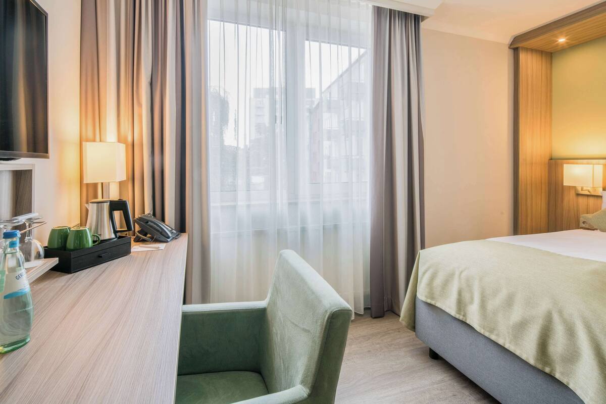 A hotel room with a bed, a desk with a telephone, a chair, and a window with curtains.
