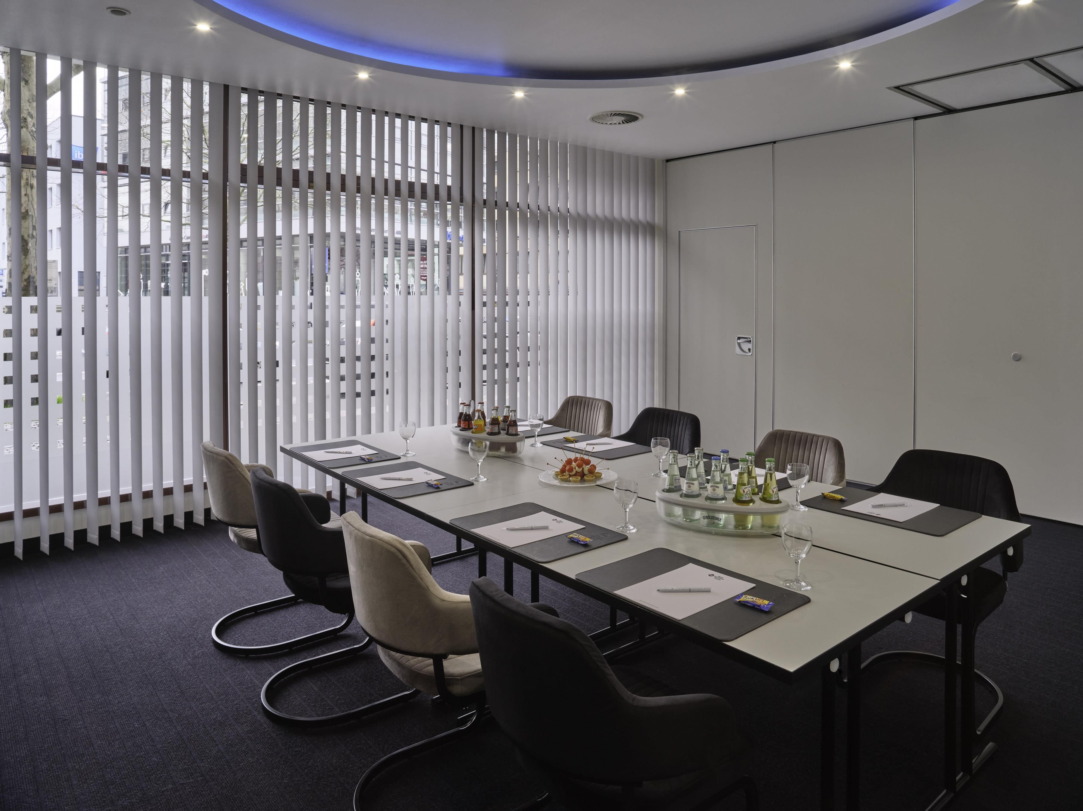 Ein Konferenzraum mit einem langen Tisch, Stühlen und einer Glastrennwand mit vertikalen Jalousien.
