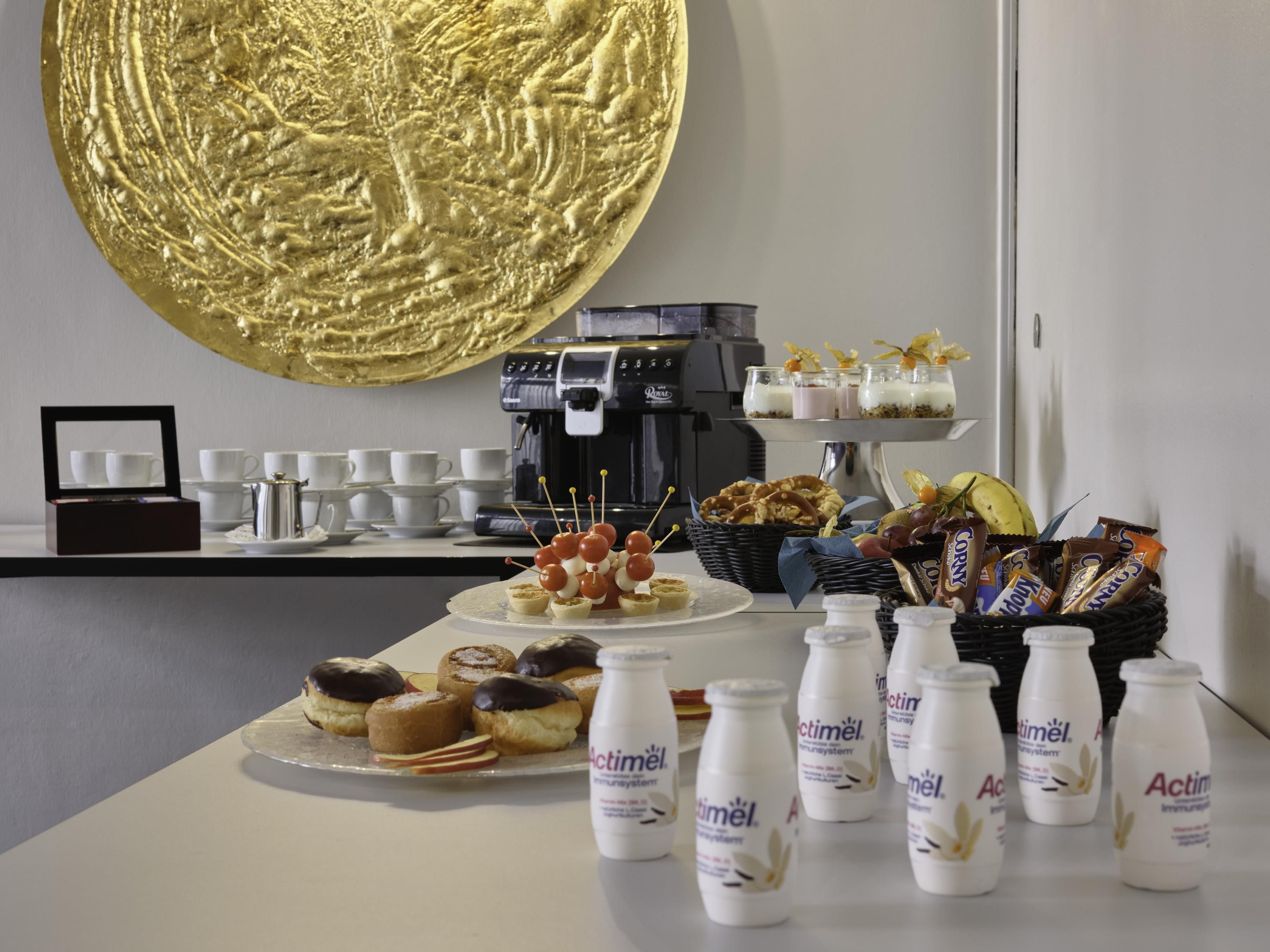 A buffet table with various food items, including pastries, a coffee machine, and bottles of Actimel.