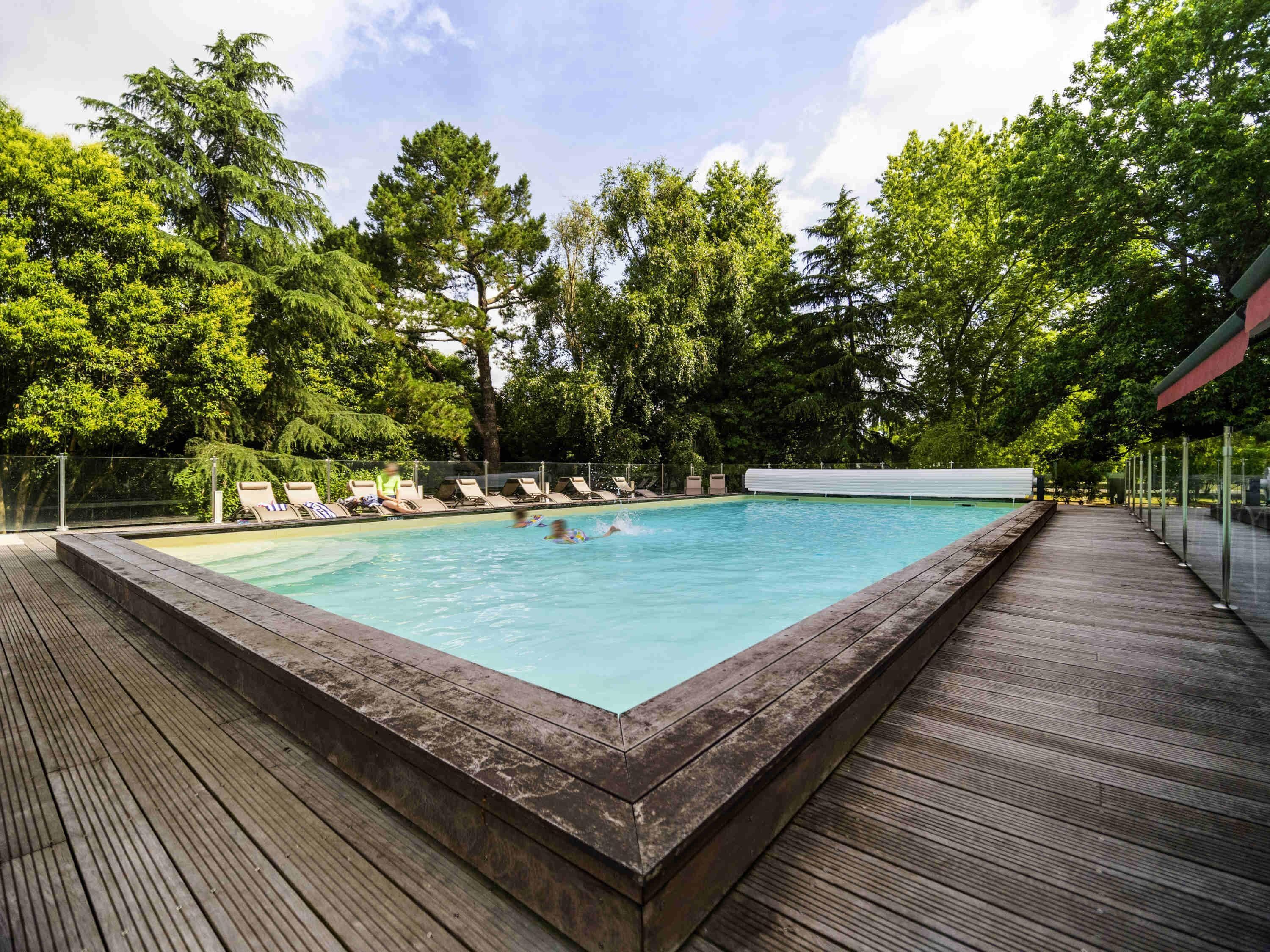 Una piscina rettangolare con un pontile in legno che la circonda, immersa nel verde degli alberi.