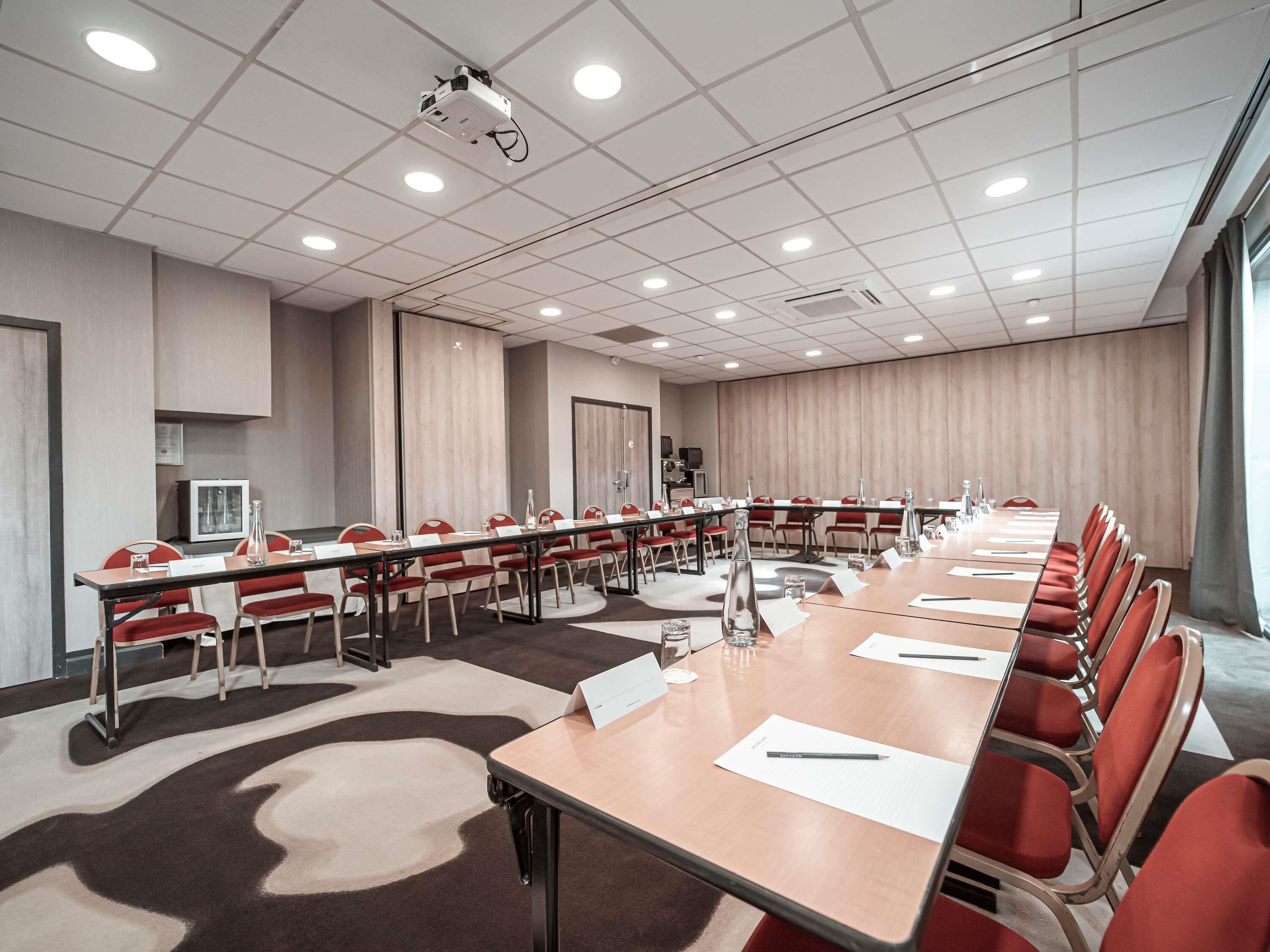 Une salle de conférence avec une longue table, des chaises rouges, des plaques nominatives et un projecteur.