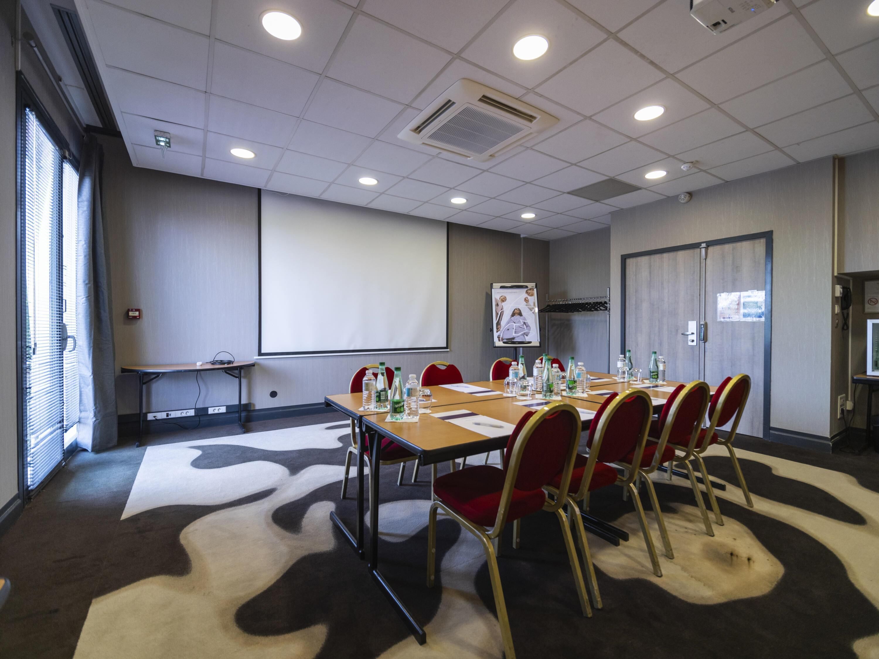 Una sala de conferencias con una pantalla grande, un proyector, una mesa con sillas y una alfombra estampada.
