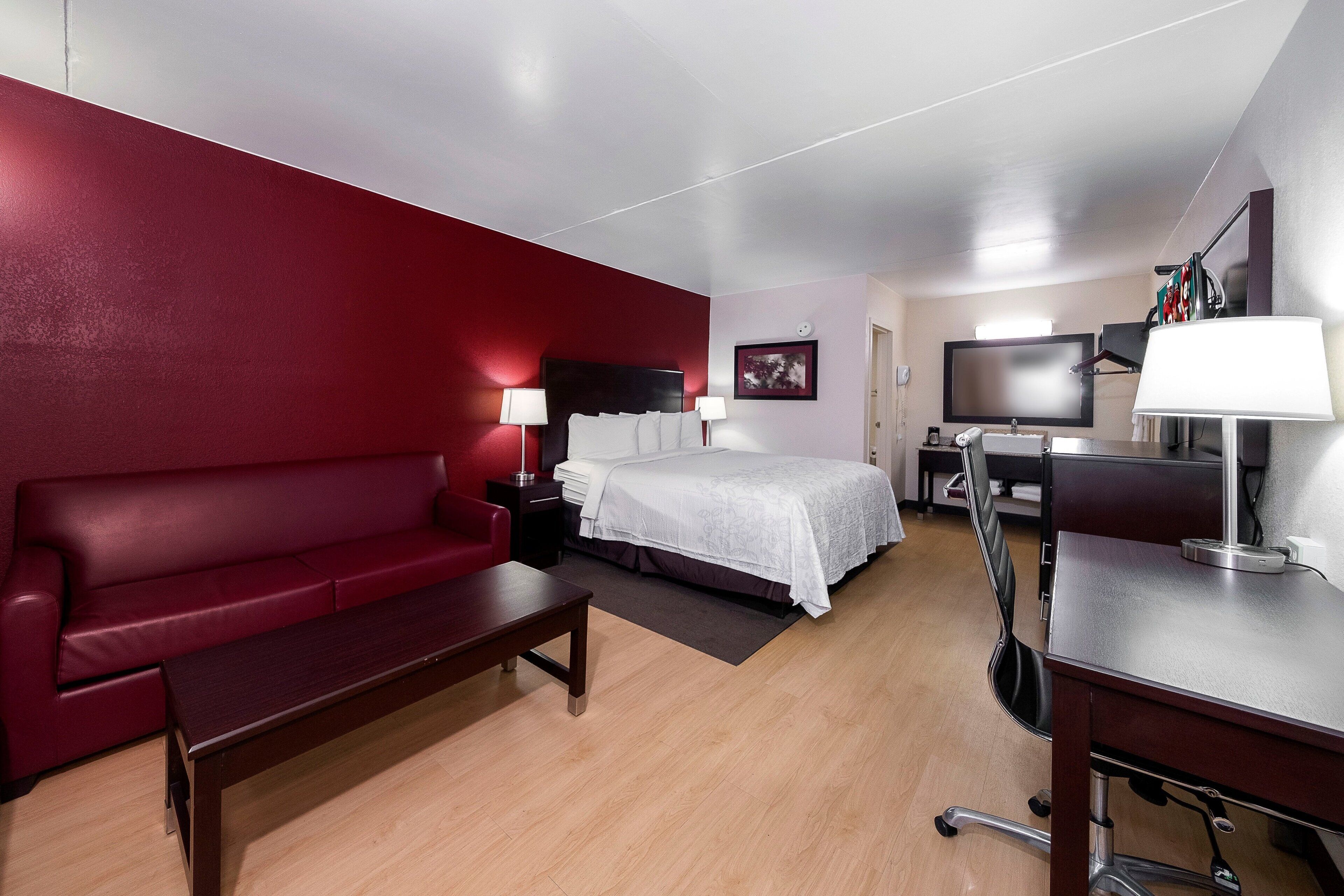 A hotel room with a red wall, a bed with white bedding, a desk with a computer, a TV, and a lamp.