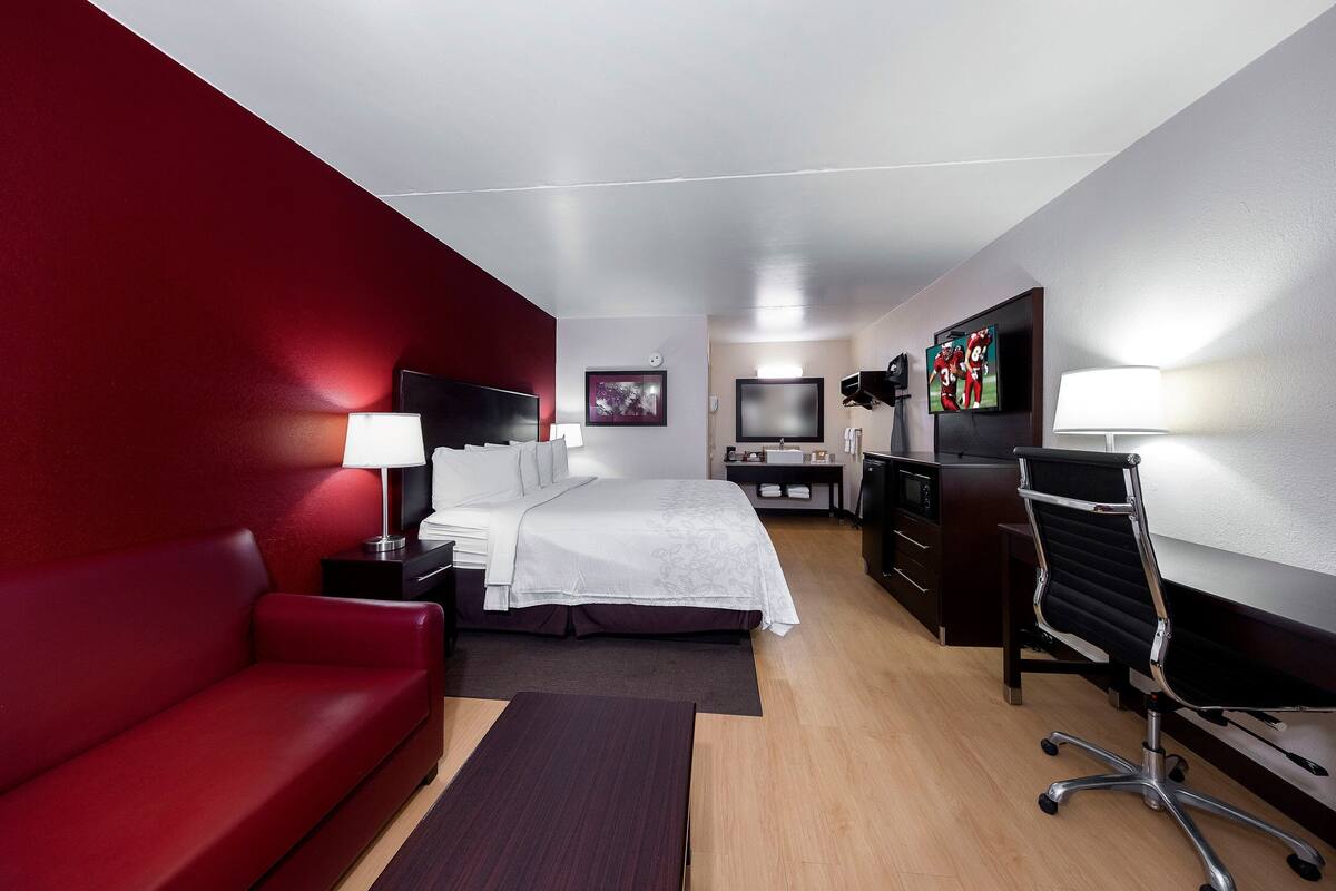 Une chambre d’hôtel avec un mur rouge, un lit avec une literie blanche, un bureau avec une chaise, une télévision et une table de chevet avec une lampe.