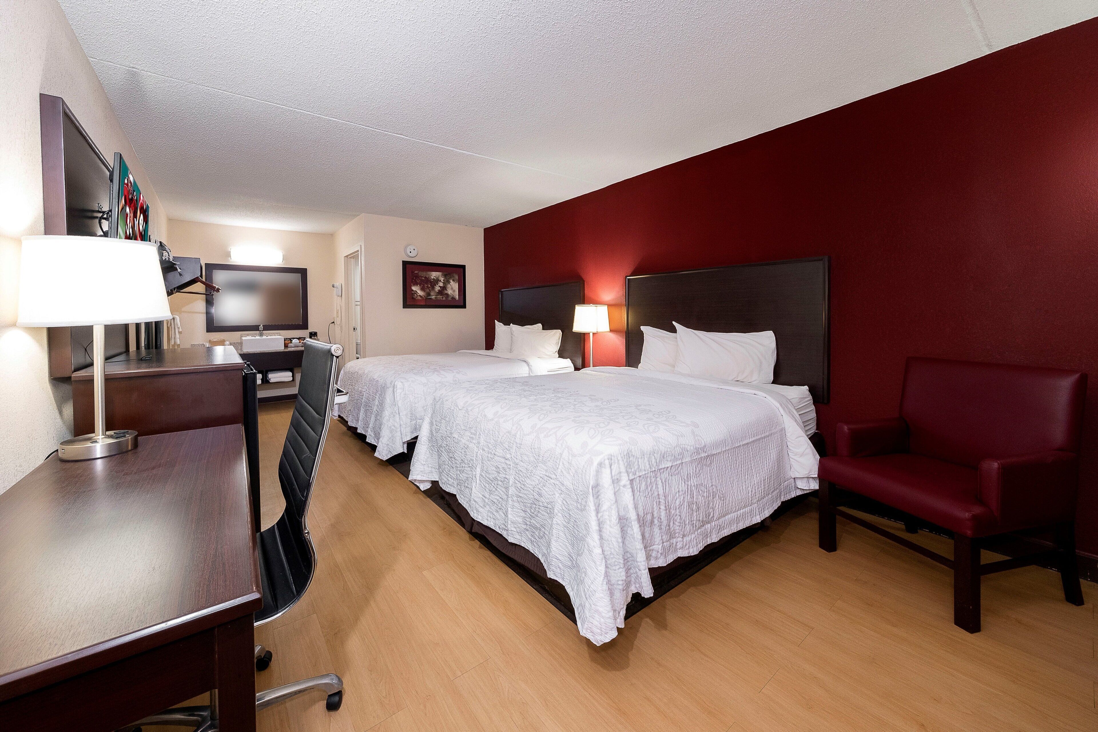 A hotel room with two beds, a desk, a chair, a TV, and a red accent wall.