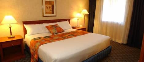 Habitación de hotel con una cama grande, dos mesitas de noche con lámparas, un cuadro enmarcado en la pared y una ventana con cortinas.