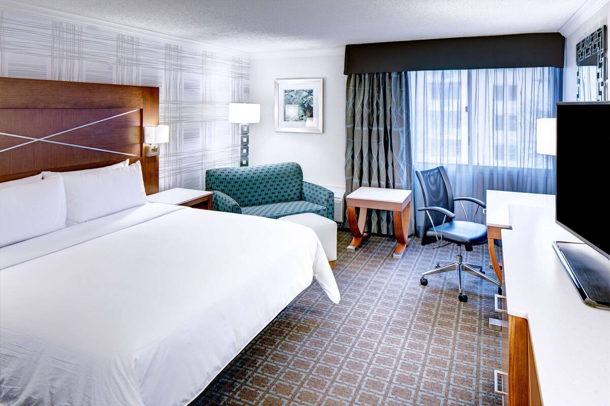 A hotel room with a large bed, a desk, a chair, a TV, and a window with curtains.