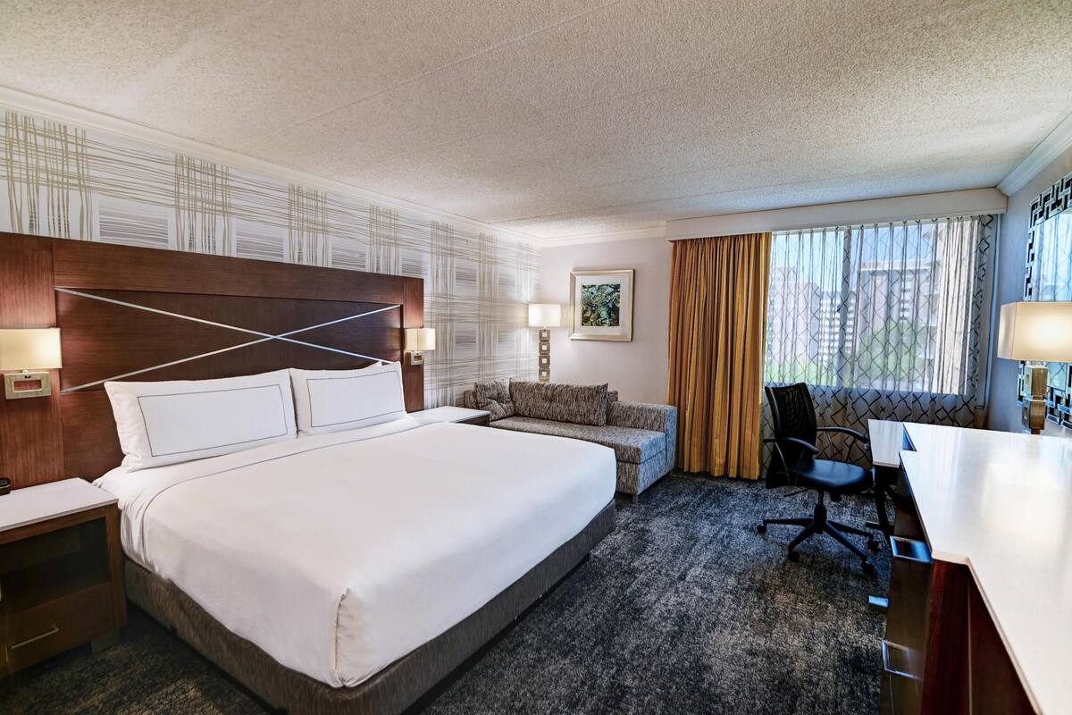 A hotel room with a large bed, a desk, a chair, a sofa, and a window with curtains.
