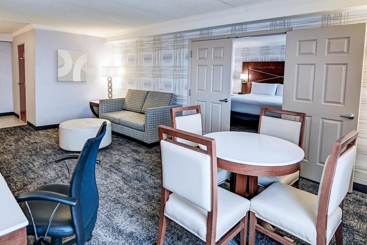 A hotel room with a round table, two chairs, a sofa, a small ottoman, a desk, and a bed with a headboard.