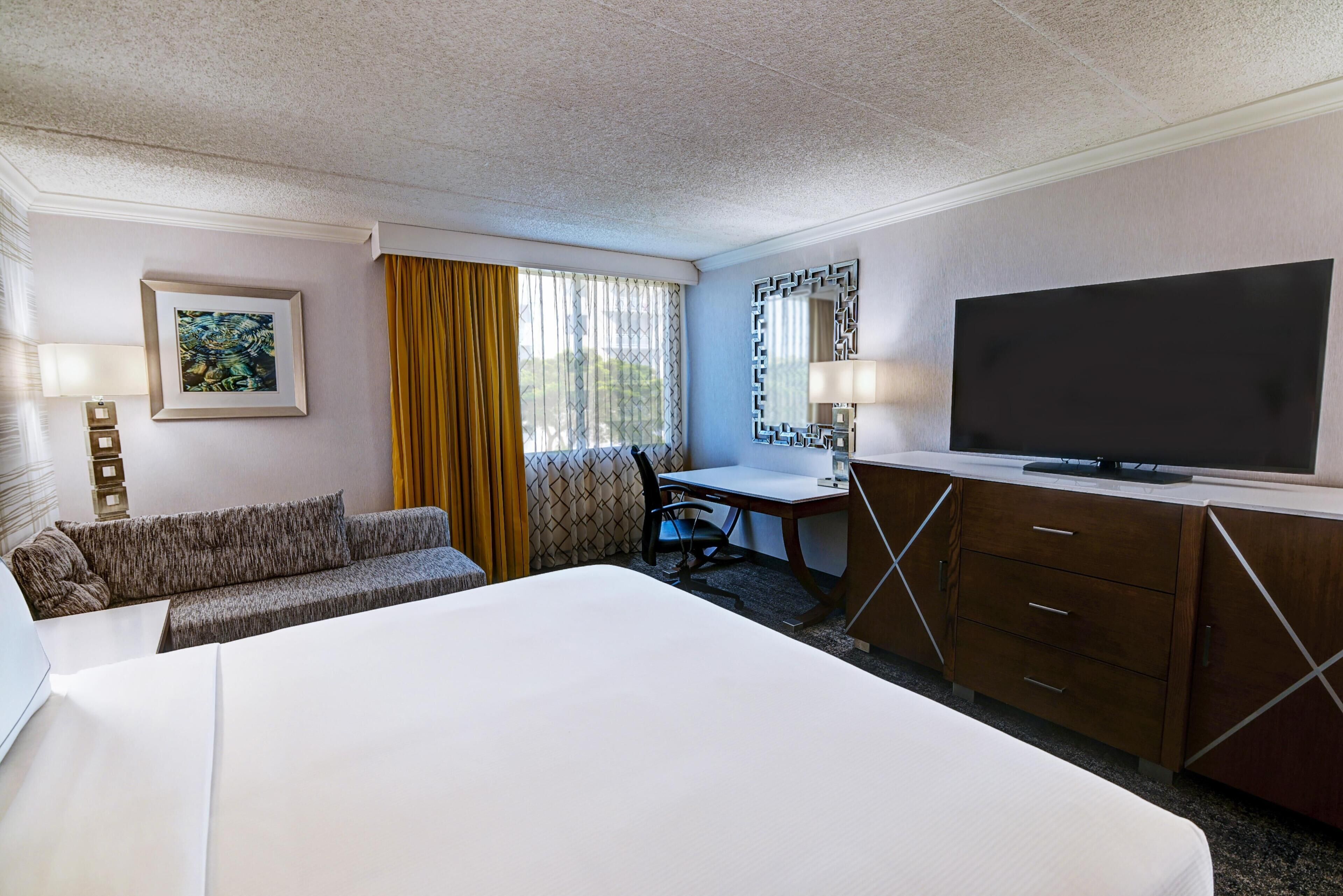 A hotel room with a large bed, a desk with a chair, a television, a sofa, and a window with curtains.