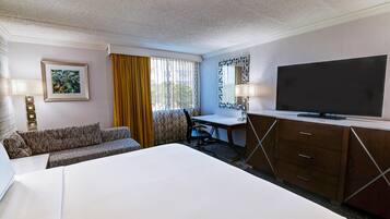A hotel room with a large bed, a desk with a chair, a television, a sofa, and a window with curtains.