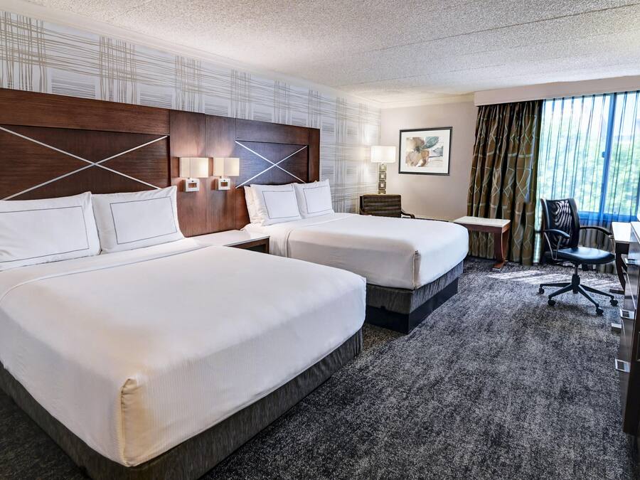 Una habitación de hotel con dos camas, un escritorio, una silla y un ventanal con cortinas.